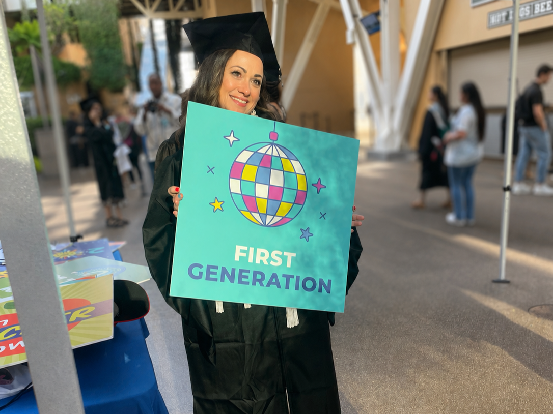 Happy woman in a graduation cap and gown holding a sign that says 'First Generation' with a colorful disco ball graphic, outdoors at graduation event.