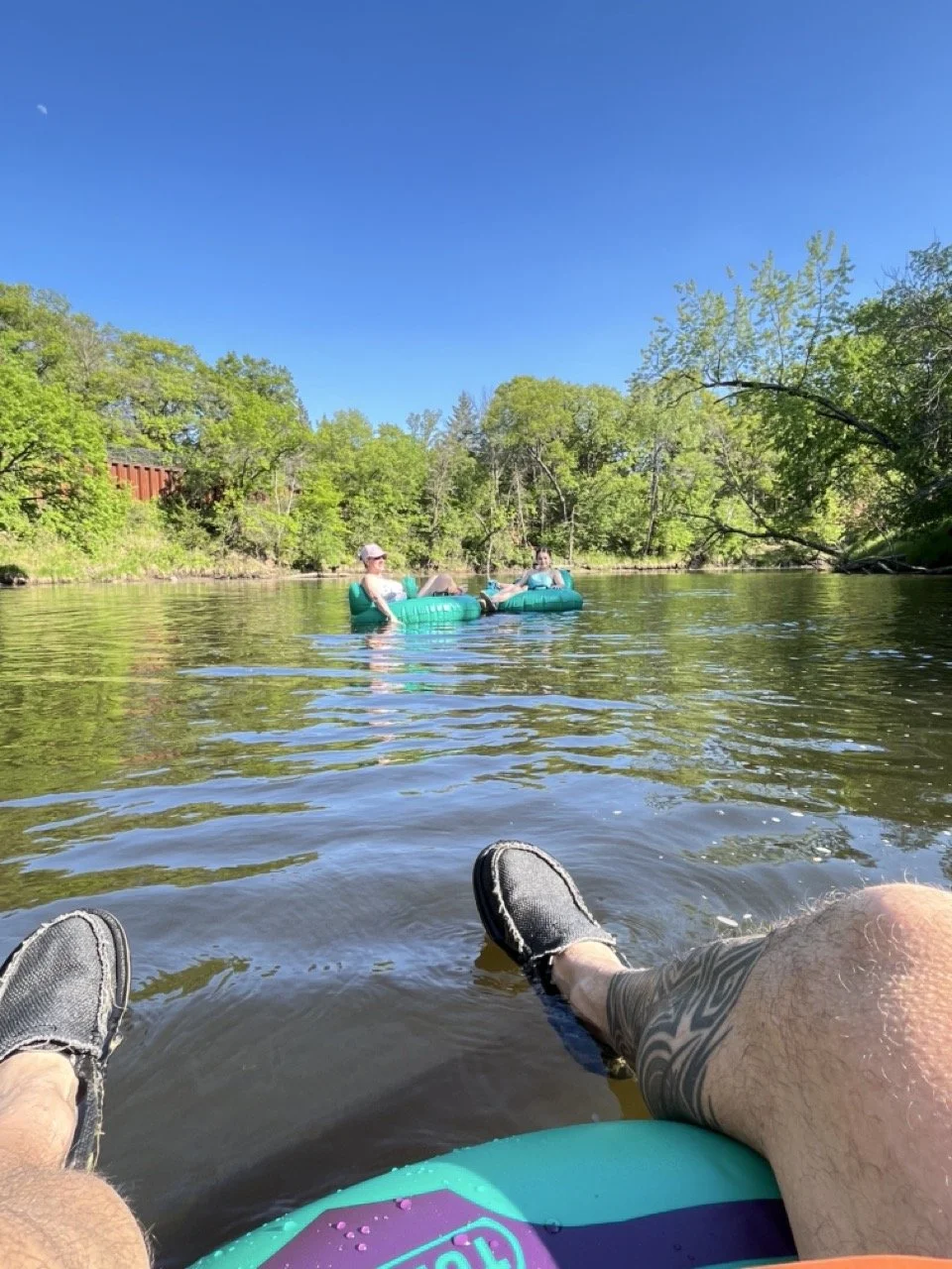People floating on inflatable tubes in a river under a clear blue sky, with green trees along the riverbank and a person's legs in the foreground.