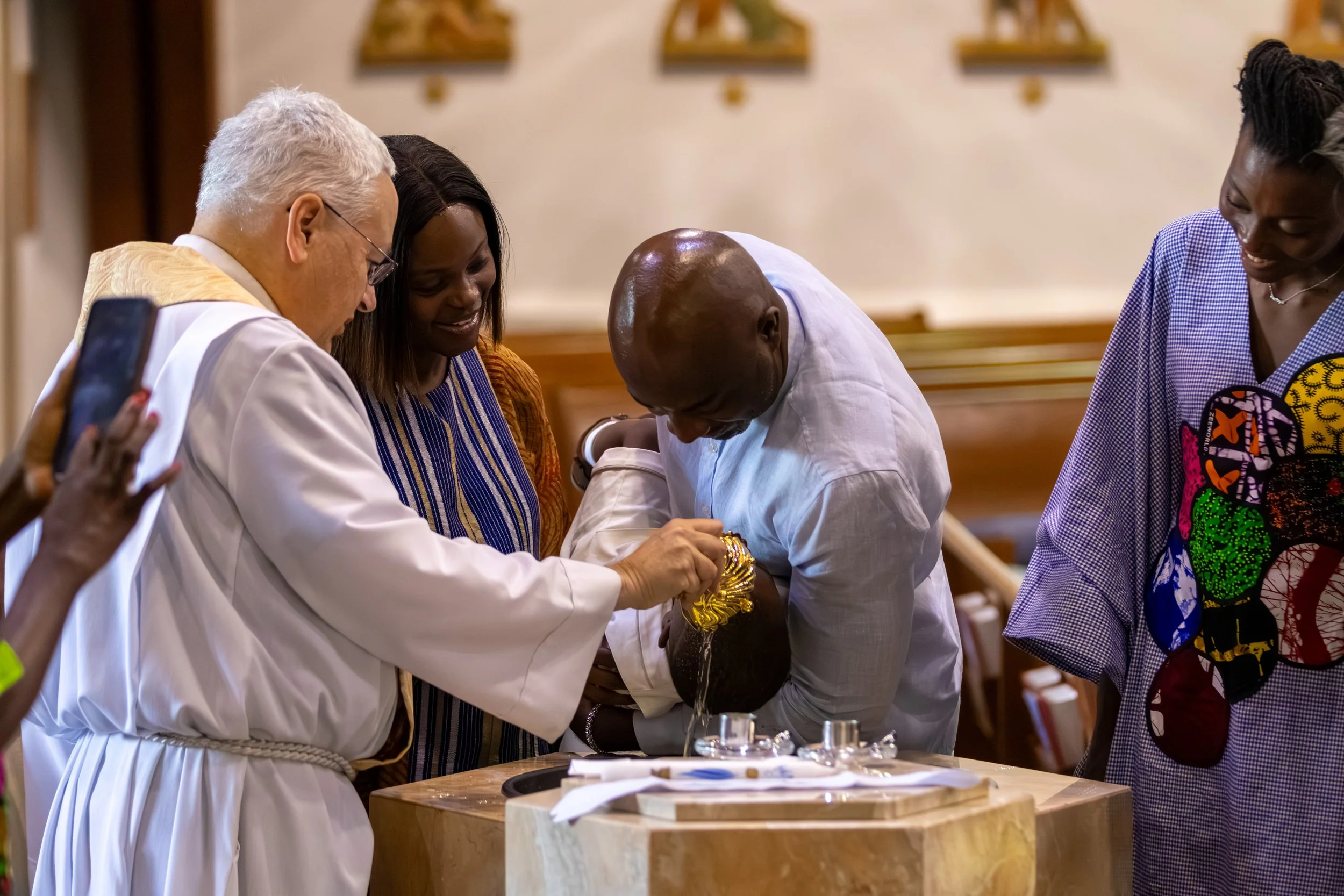 A religious ceremony taking place in a church where a man is receiving a blessing from a priest while others look on smiling. The priest is wearing a white robe with a golden stole, and the setting includes wooden pews and religious artwork on the wall.