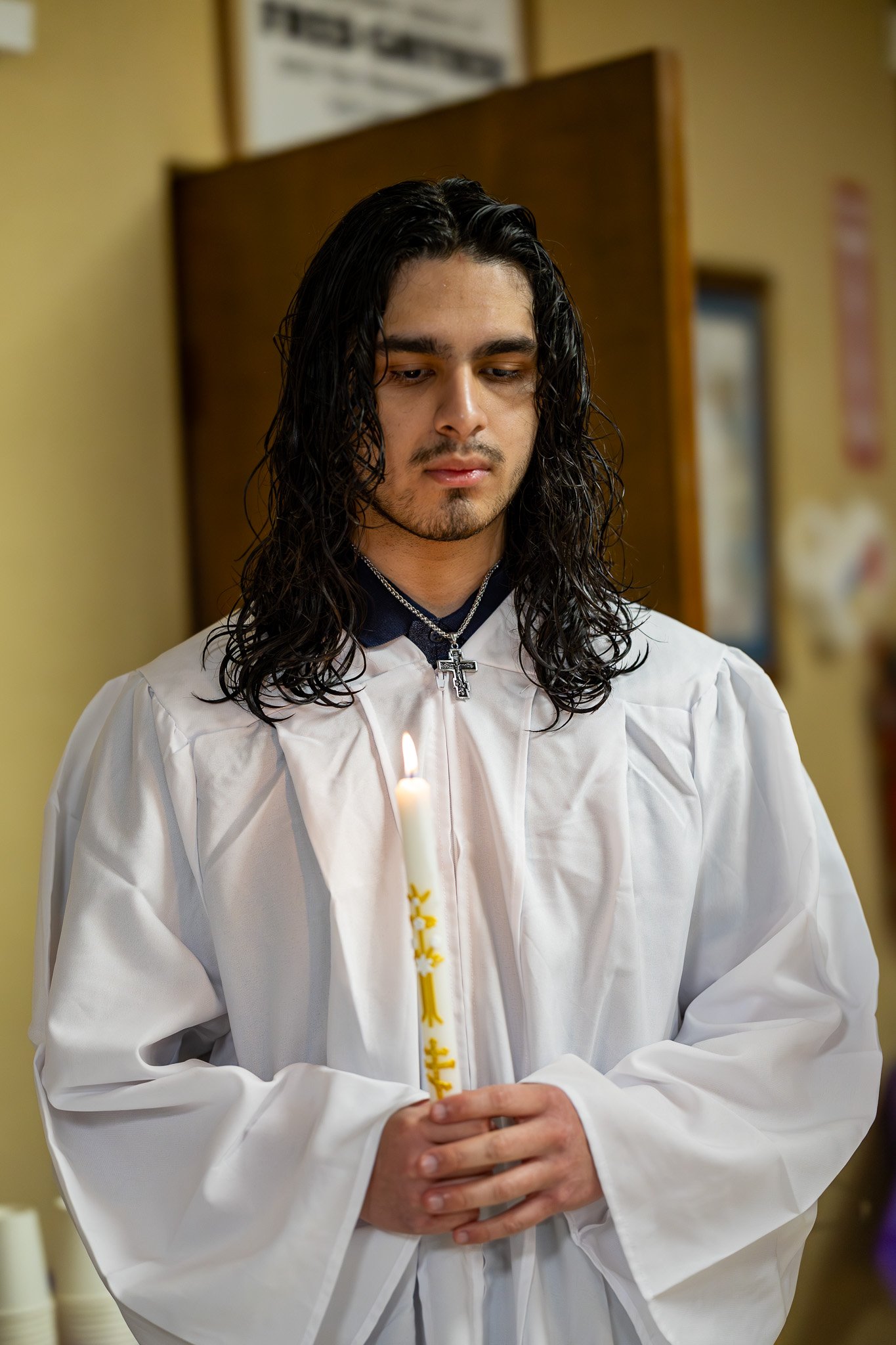 A young man with long, dark curly hair wearing a white religious robe and a silver cross necklace, holding a lit candle during a religious ceremony.