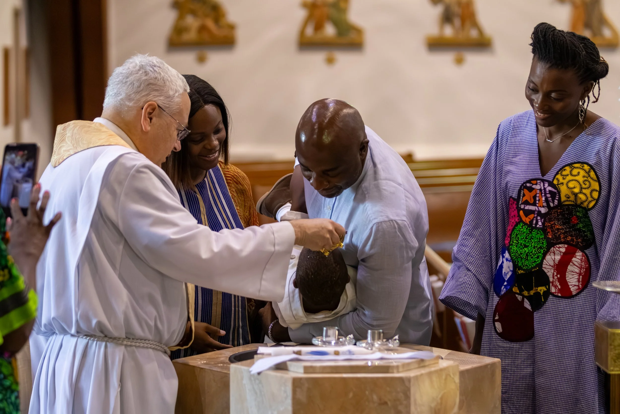 A group of people, including a priest, gathered around a baptismal font during a baptism ceremony in a church, with a priest pouring water on a child's head.