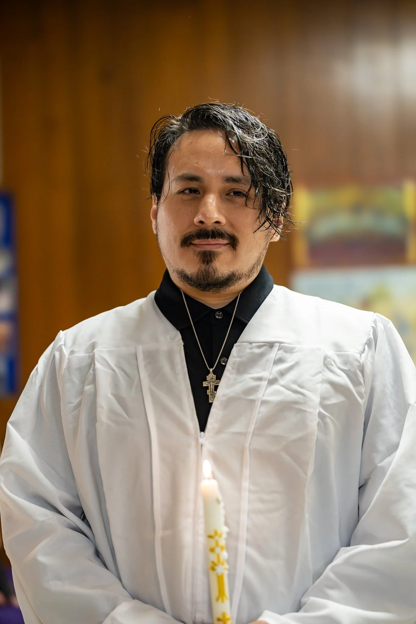 A man dressed in a white robe with a black collar and a silver cross necklace, standing in front of a blurred background, holding a lit candle with a decorative yellow and white pattern.