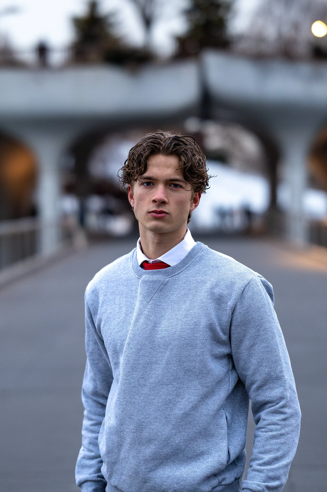 Young man with curly brown hair standing outdoors on a street, wearing a light gray sweater over a collared shirt and red tie, with blurred background of trees and buildings.