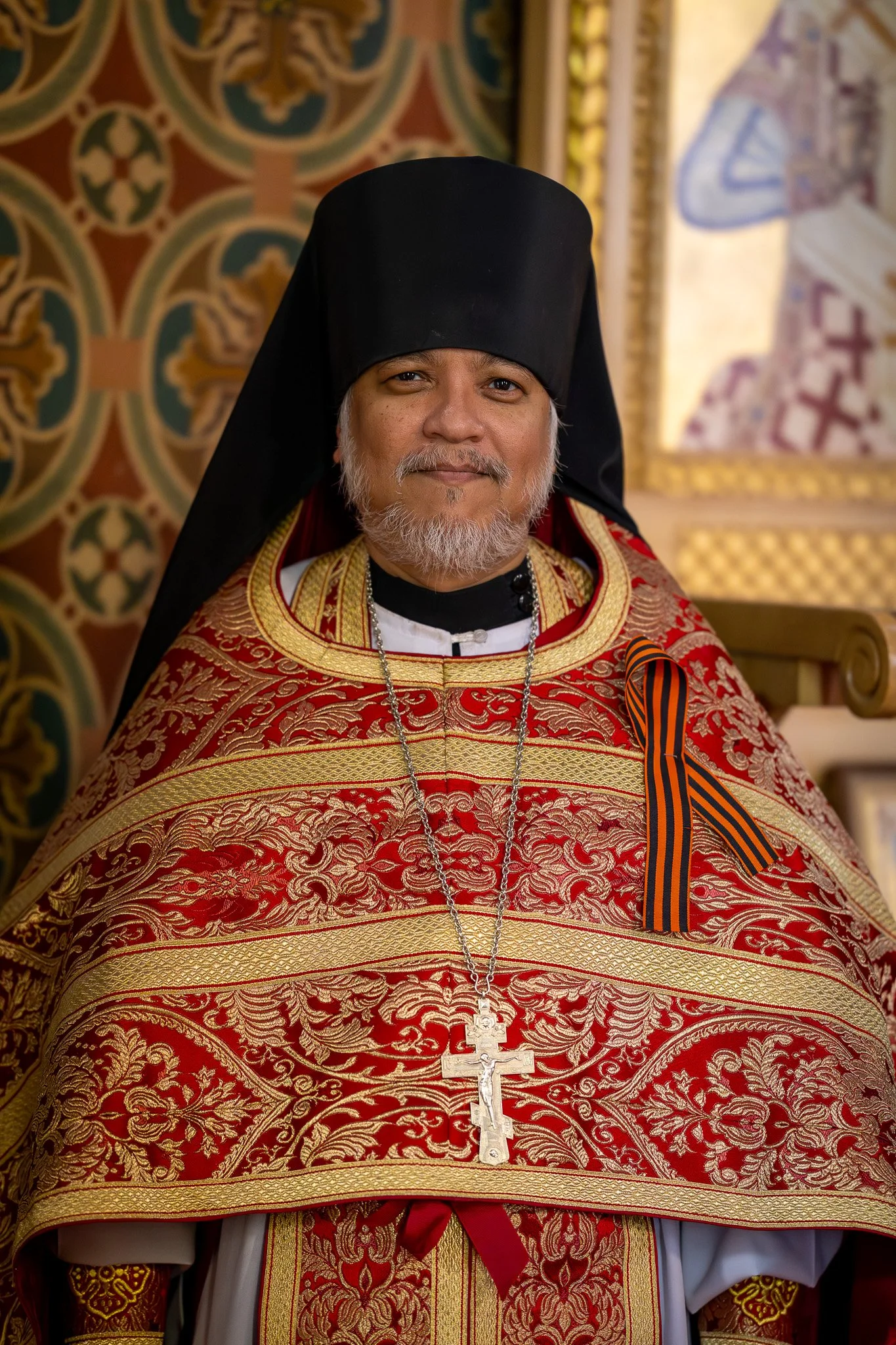 Man dressed in ornate religious robes with gold embroidery, wearing a black hat, gold and red necklace, and Jesus cross pendant, with a patterned background.