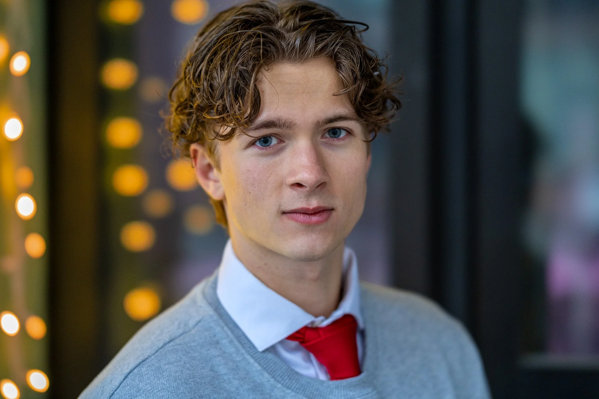 A young man with curly brown hair and blue eyes wearing a light gray blazer, white shirt, and red tie, standing indoors near a background with blurred yellow lights.