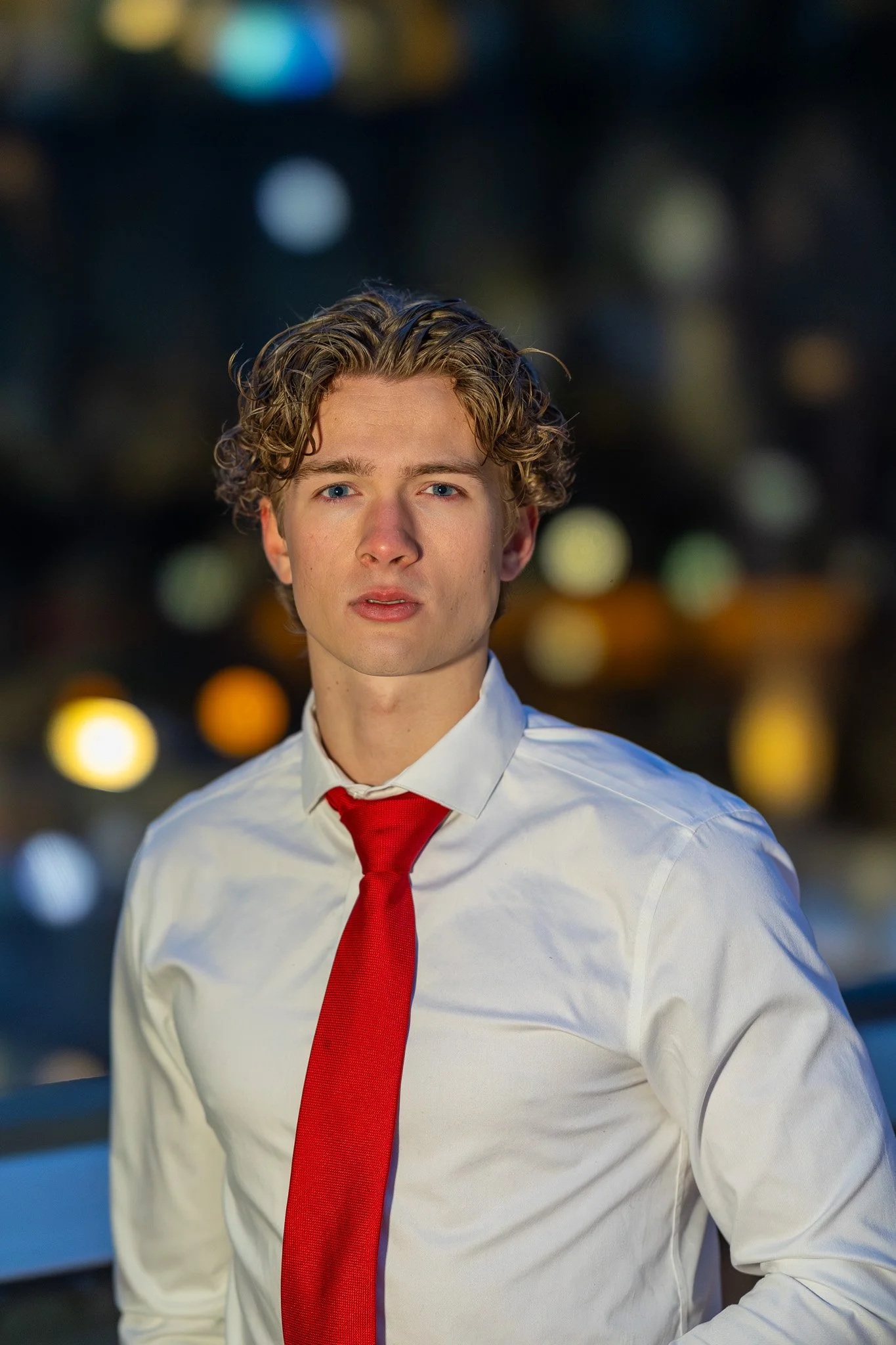 A young man with curly blond hair looking directly at the camera, wearing a white shirt and a red tie, with a blurred cityscape background at night.