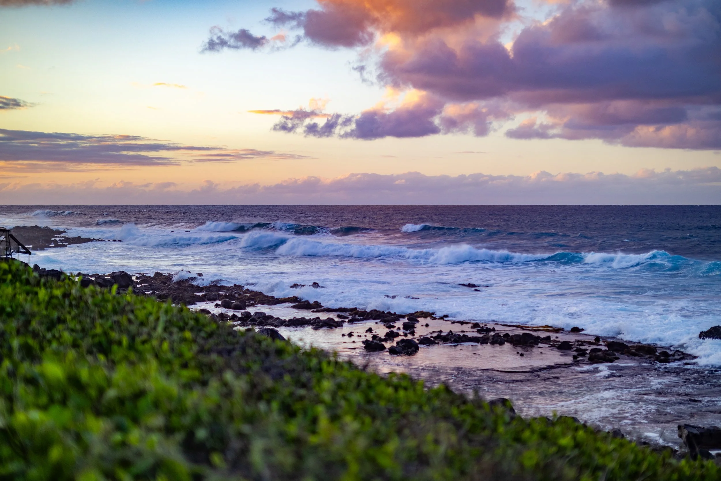 Sunset over the ocean with waves crashing onto rocky shoreline and colorful clouds in the sky.