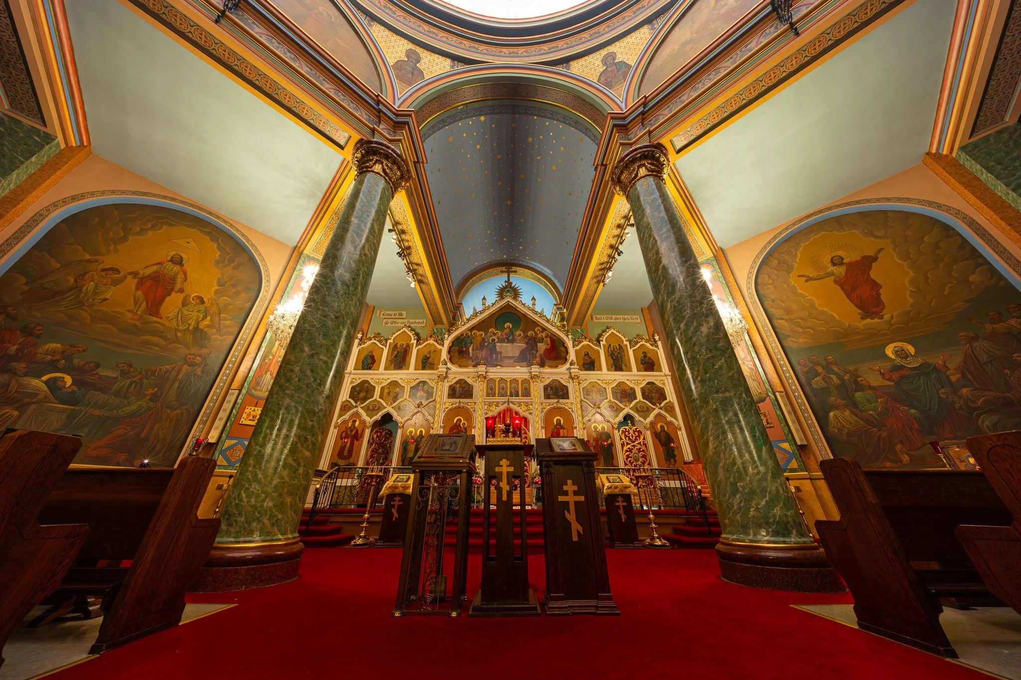 Interior of an Orthodox church with ornate icons, gold accents, tall green marble columns, and a red carpet leading to the altar.