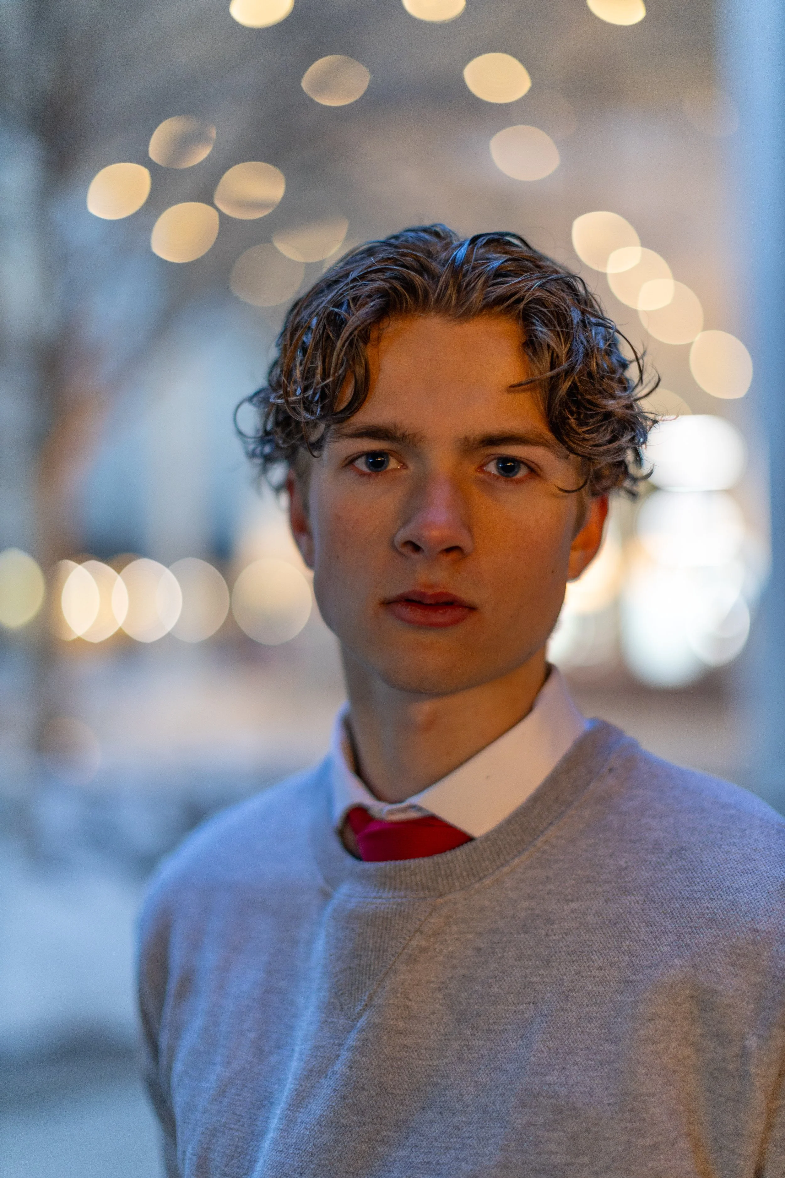 Close-up of a young man with wavy brown hair, blue eyes, wearing a light gray sweater, white shirt, and red tie, standing outdoors at dusk with blurred city lights in the background.