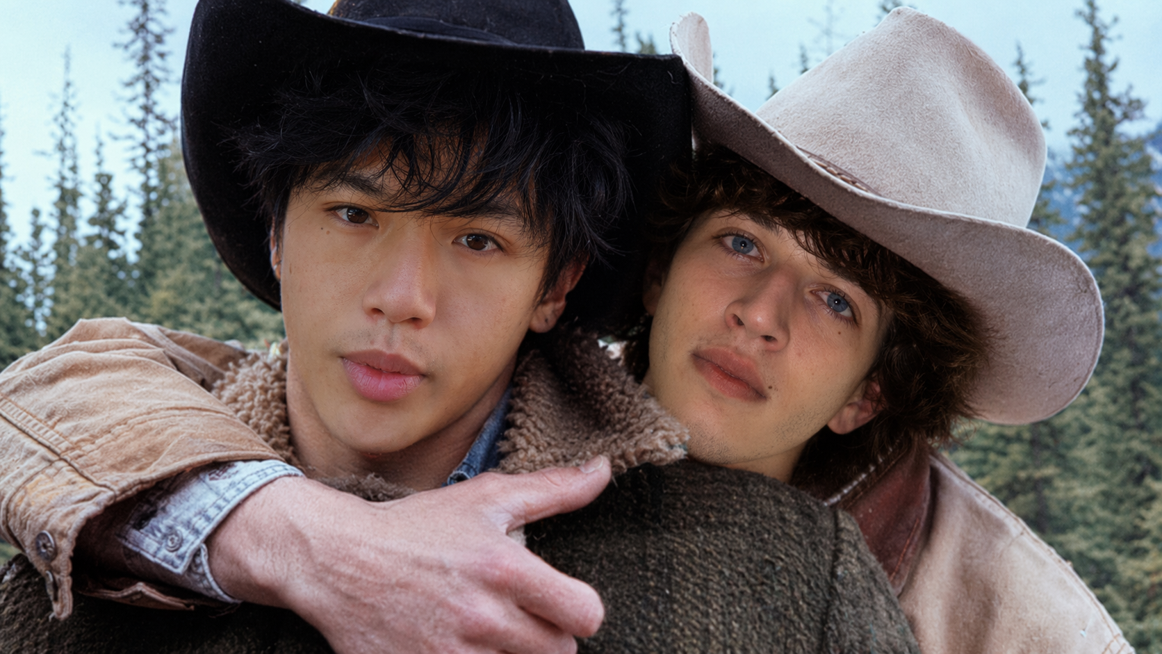 Two young men wearing cowboy hats outdoors, close-up shot, with a forest in the background.