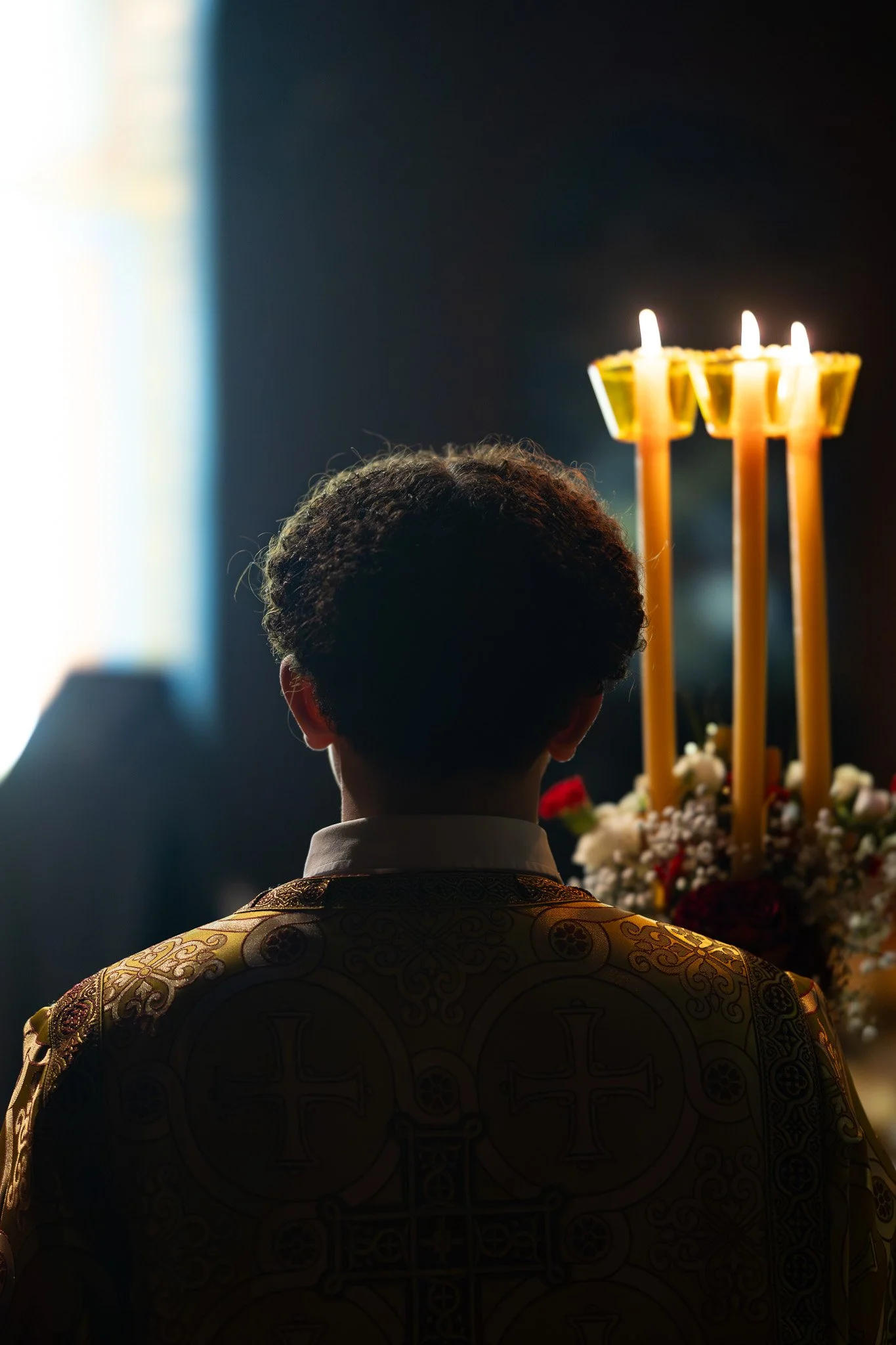 A person in traditional attire stands in front of a religious or ceremonial altar with three lit candles and flowers, illuminated by light coming from a window or opening behind them.