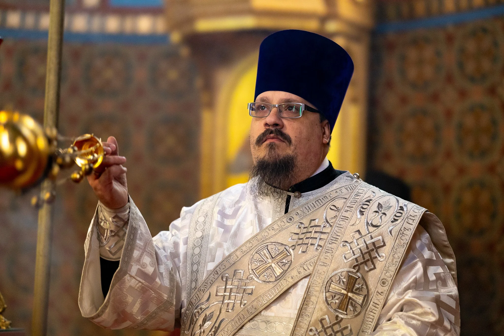 A priest wearing a dark blue mitre and glasses, holding a golden staff, dressed in ornate white and gold ceremonial robes, inside a decorated church with colorful murals on the walls.