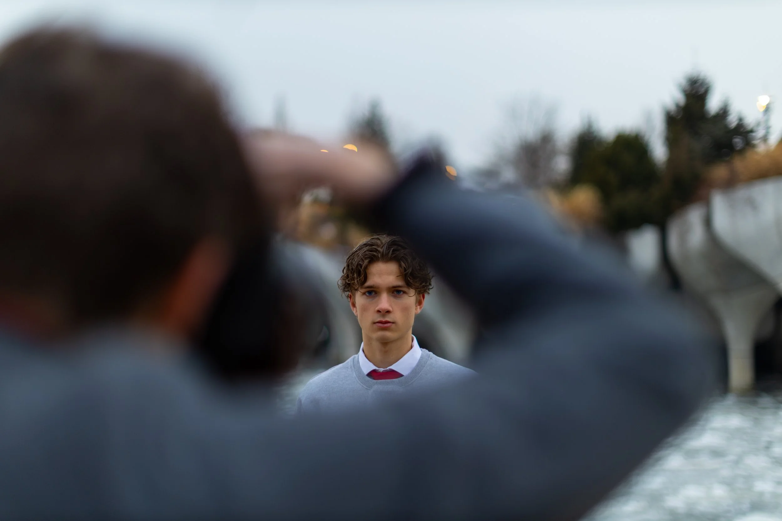 A young man with curly hair standing outdoors with trees and cloudy sky in the background, as seen through an out-of-focus foreground.