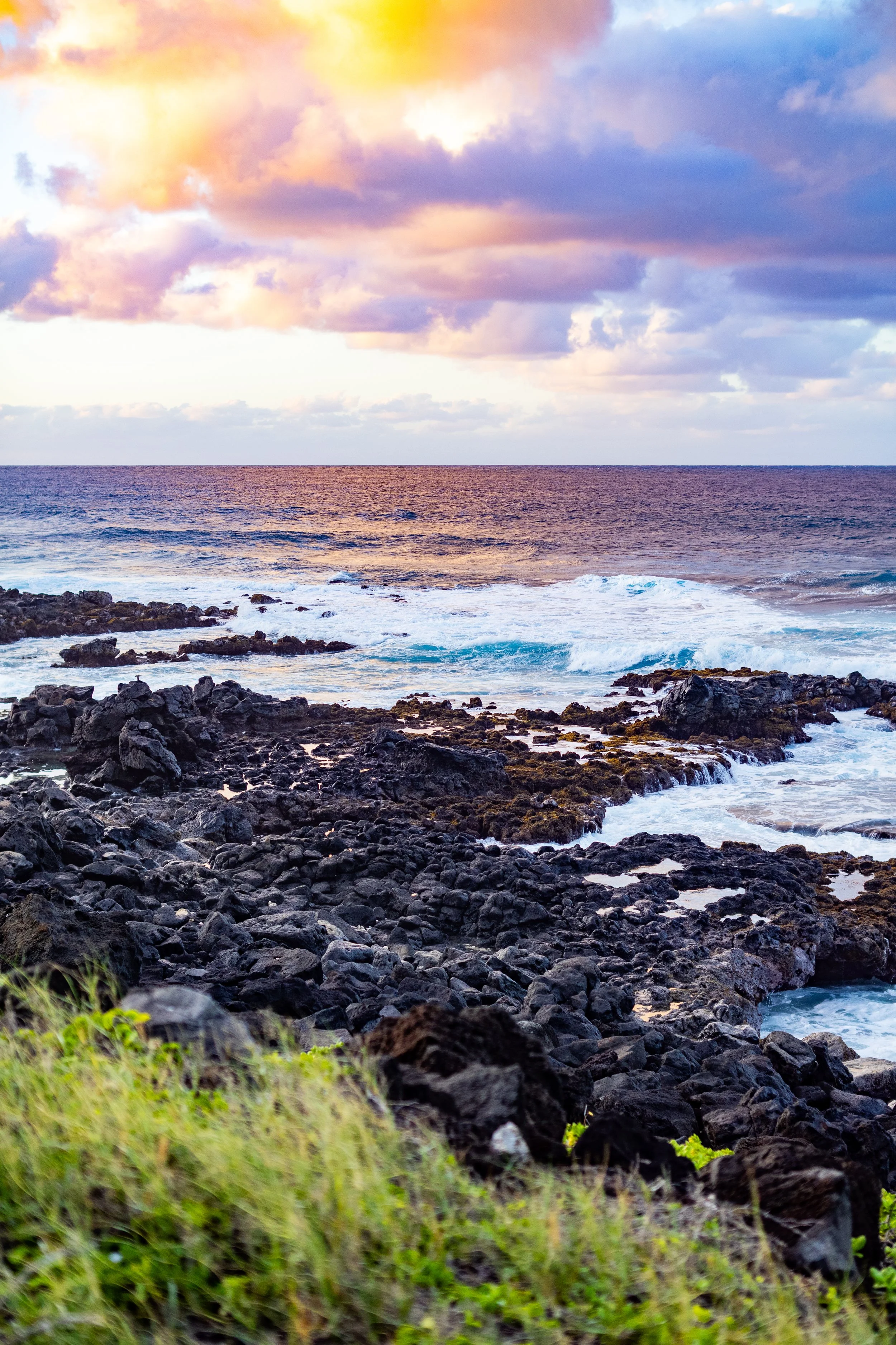 View of a rocky coastline with the ocean waves crashing against the rocks, under a sky filled with colorful clouds during sunset or sunrise.