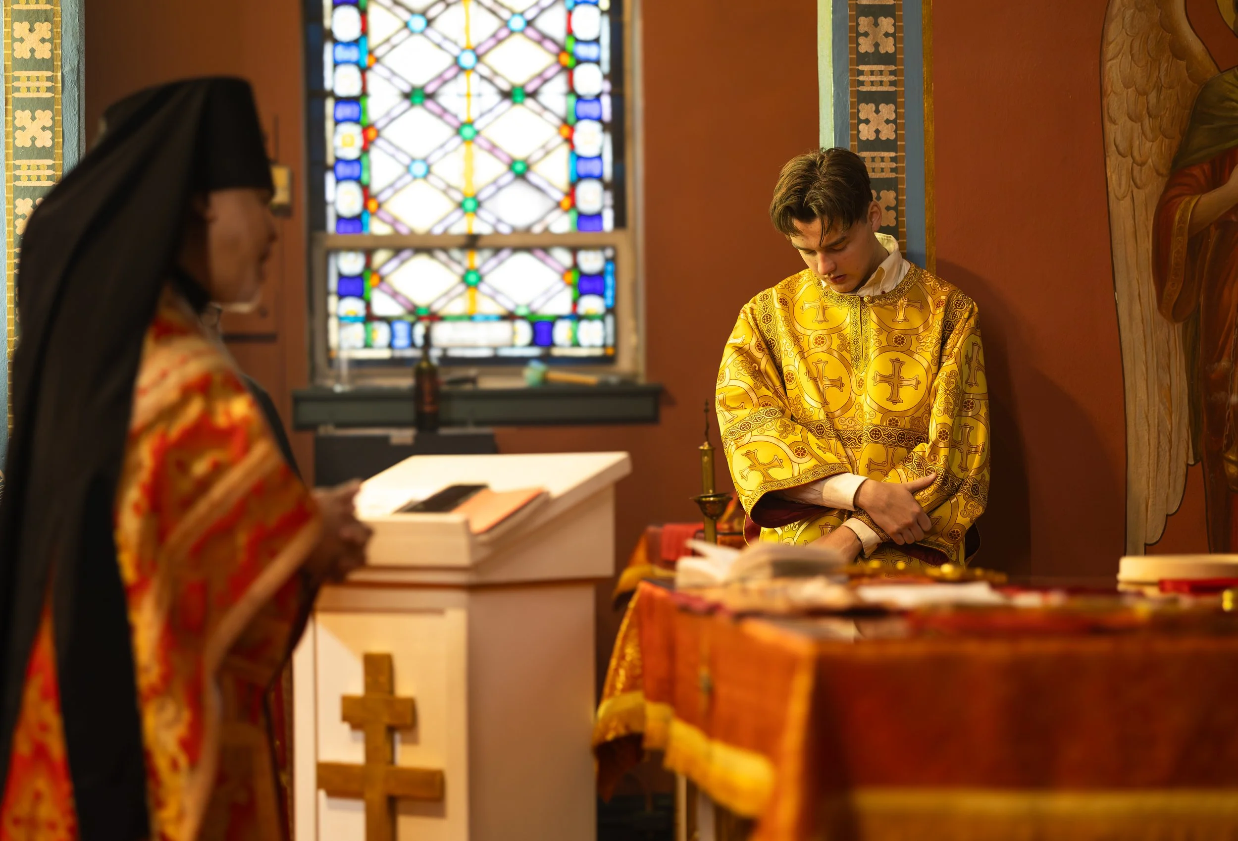 A young man and a woman in religious robes inside a church with stained glass windows. The man is bowing his head with book and cross nearby, while the woman is holding a book.
