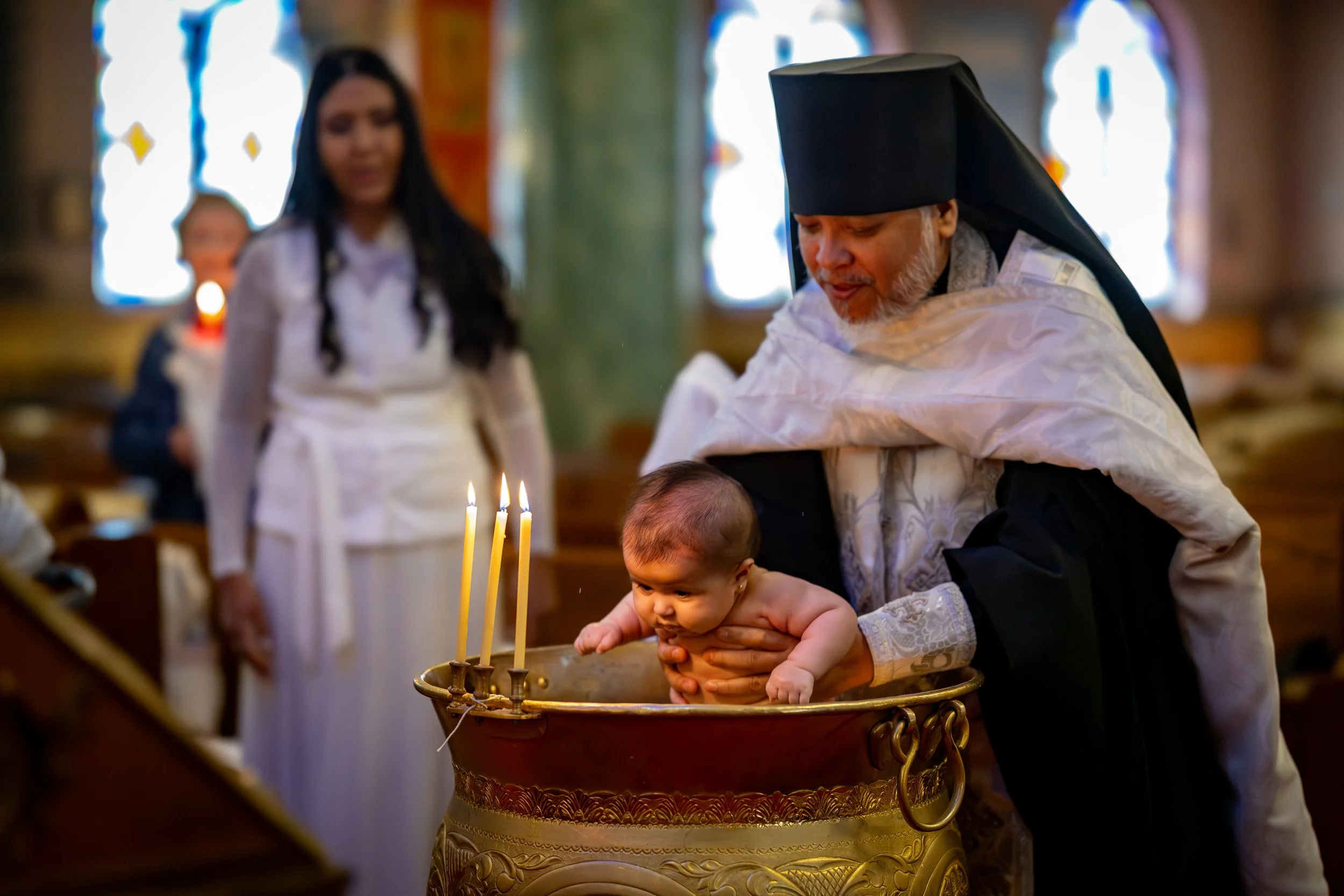 An elderly priest in traditional religious garments holds a baby over a baptismal font in a church. A woman, likely the baby's mother, stands nearby. There are lit candles and stained glass windows in the background.