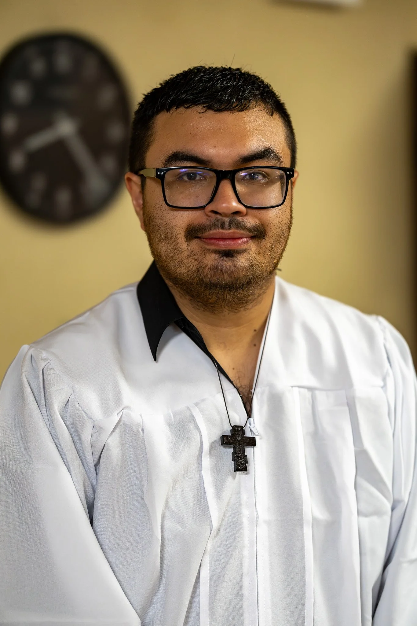 Portrait of a man wearing glasses, a white shirt with a black collar, and a black cross necklace, standing indoors with a blurred clock on the wall in the background.