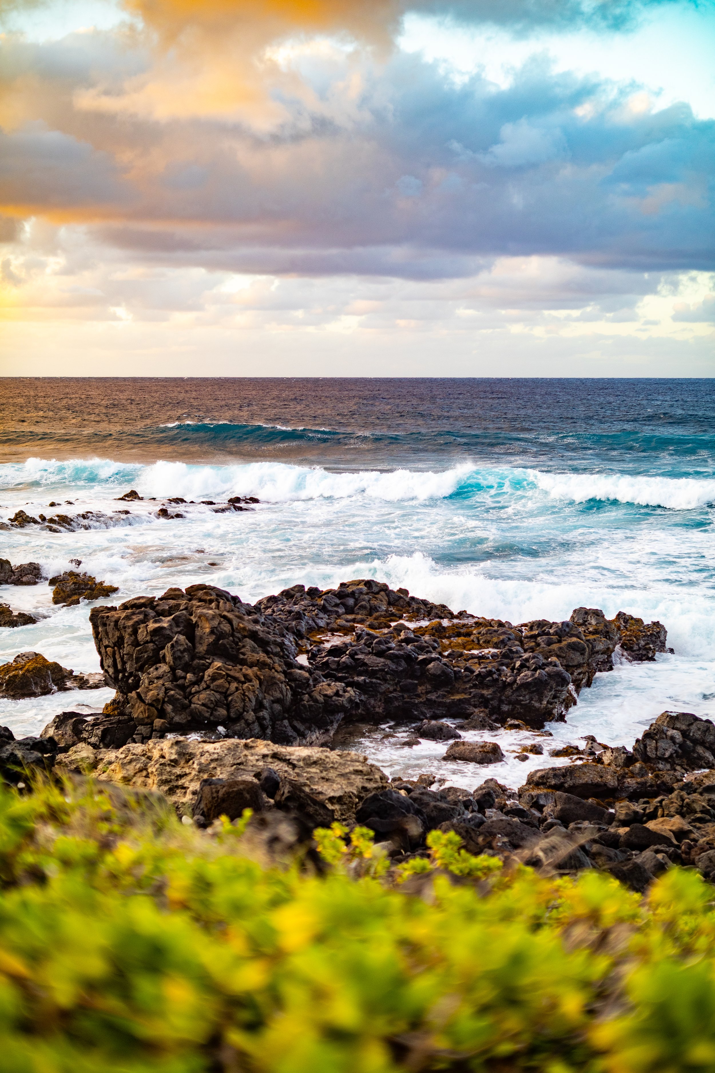 Coastline with rocks, crashing ocean waves, and a partly cloudy sky with hues of orange, blue, and purple during sunset or sunrise.