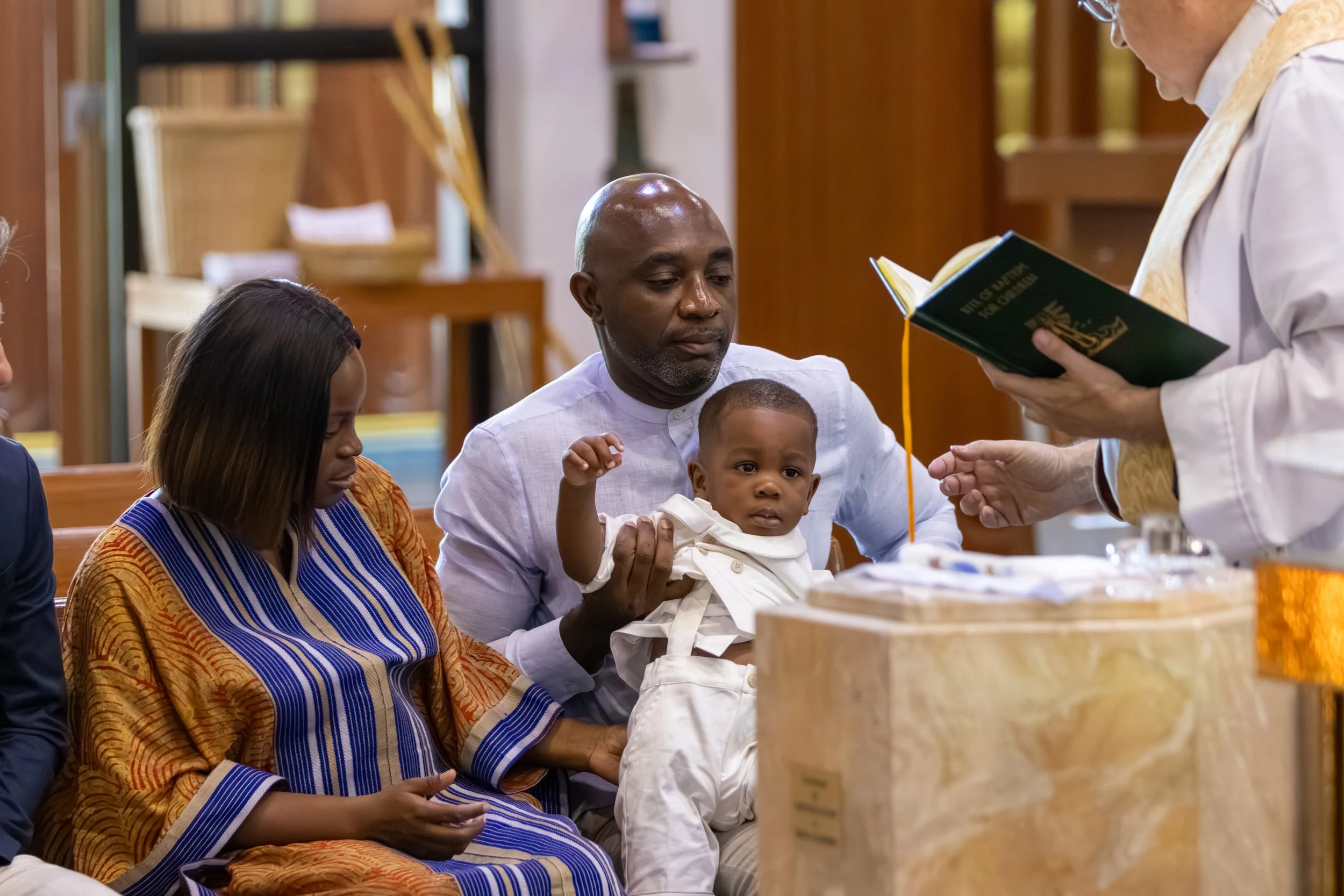 A family of four, including a man holding a young child, sits at a table during a religious service in a church. A woman in traditional African attire looks on as a person dressed in liturgical robes reads from a book.