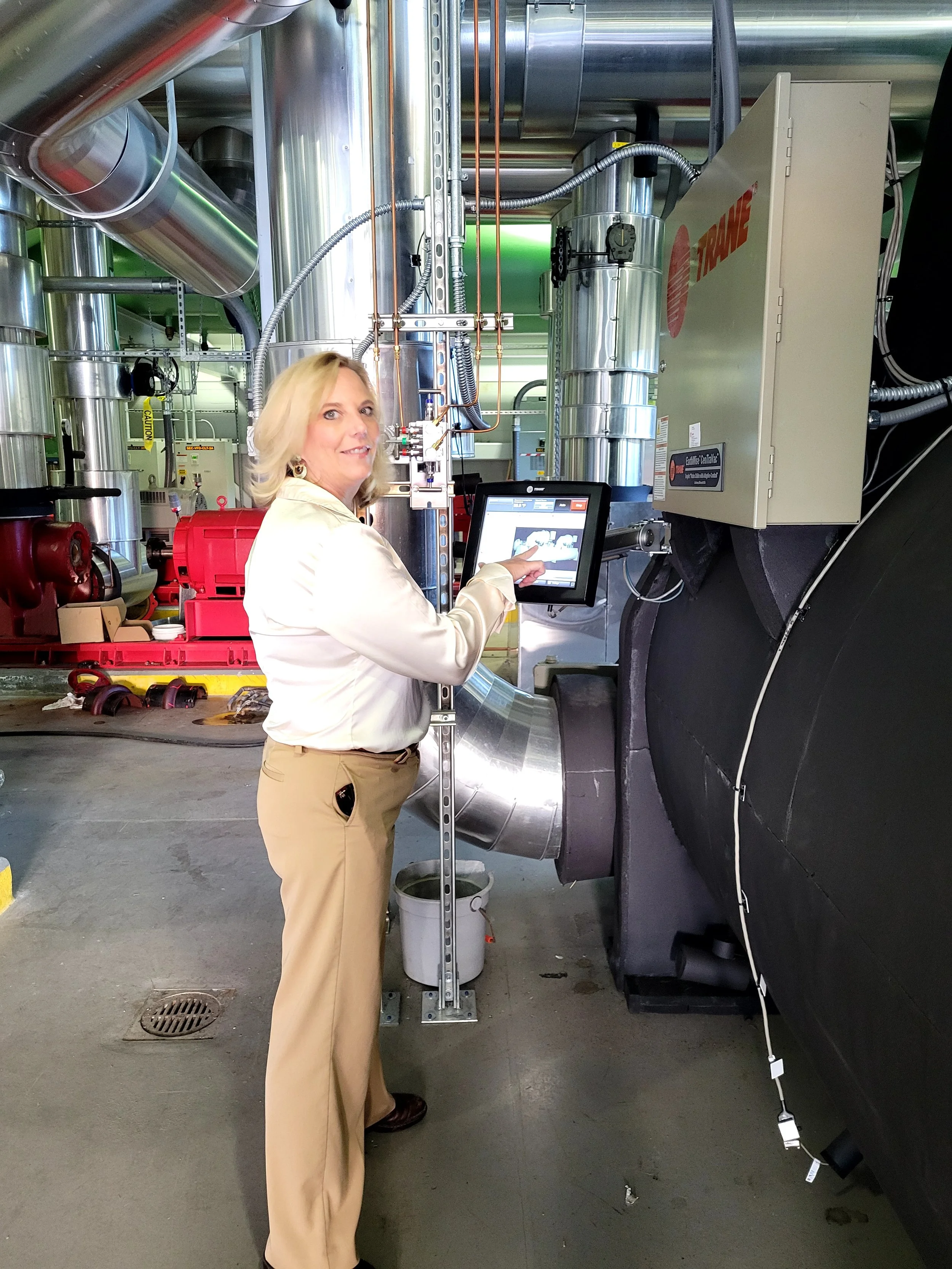 A woman with blonde hair wearing a white shirt and tan pants is standing in a or mechanical room, pointing at a computer monitor mounted on a metal stand. Behind her are large metallic pipes, tubes, and equipment, with some black insulation and a warning sign. The setting appears to be a facility related to engineering or manufacturing.
