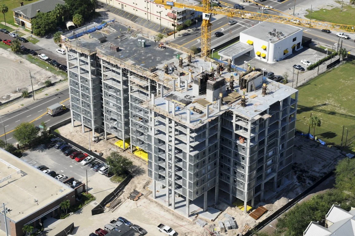 High-angle view of a multi-story building under construction with a large yellow crane on site, surrounded by parking lots, roads, and nearby buildings.