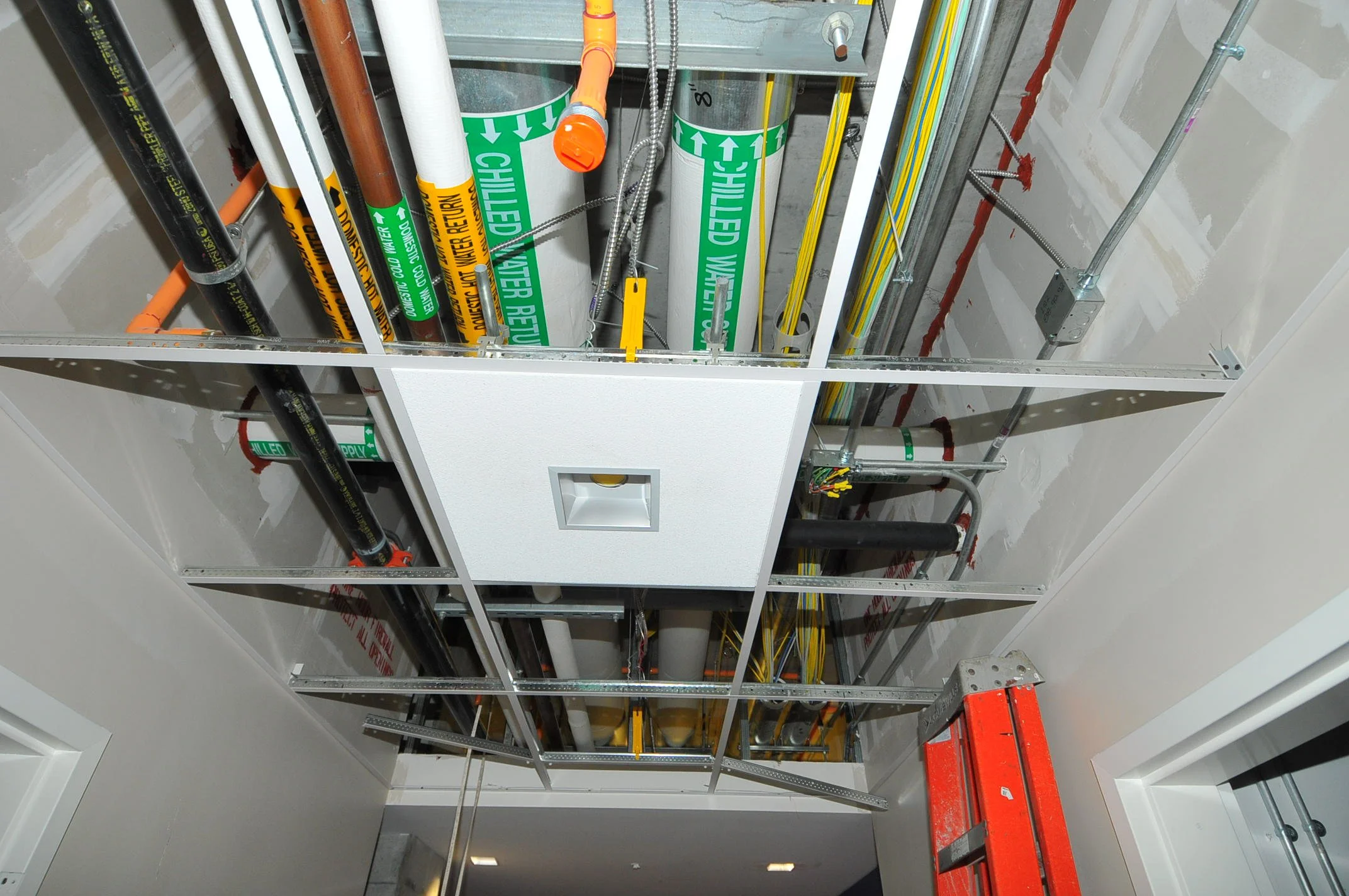View of an unfinished ceiling with exposed wiring, pipes, and metal framing in a building under construction.