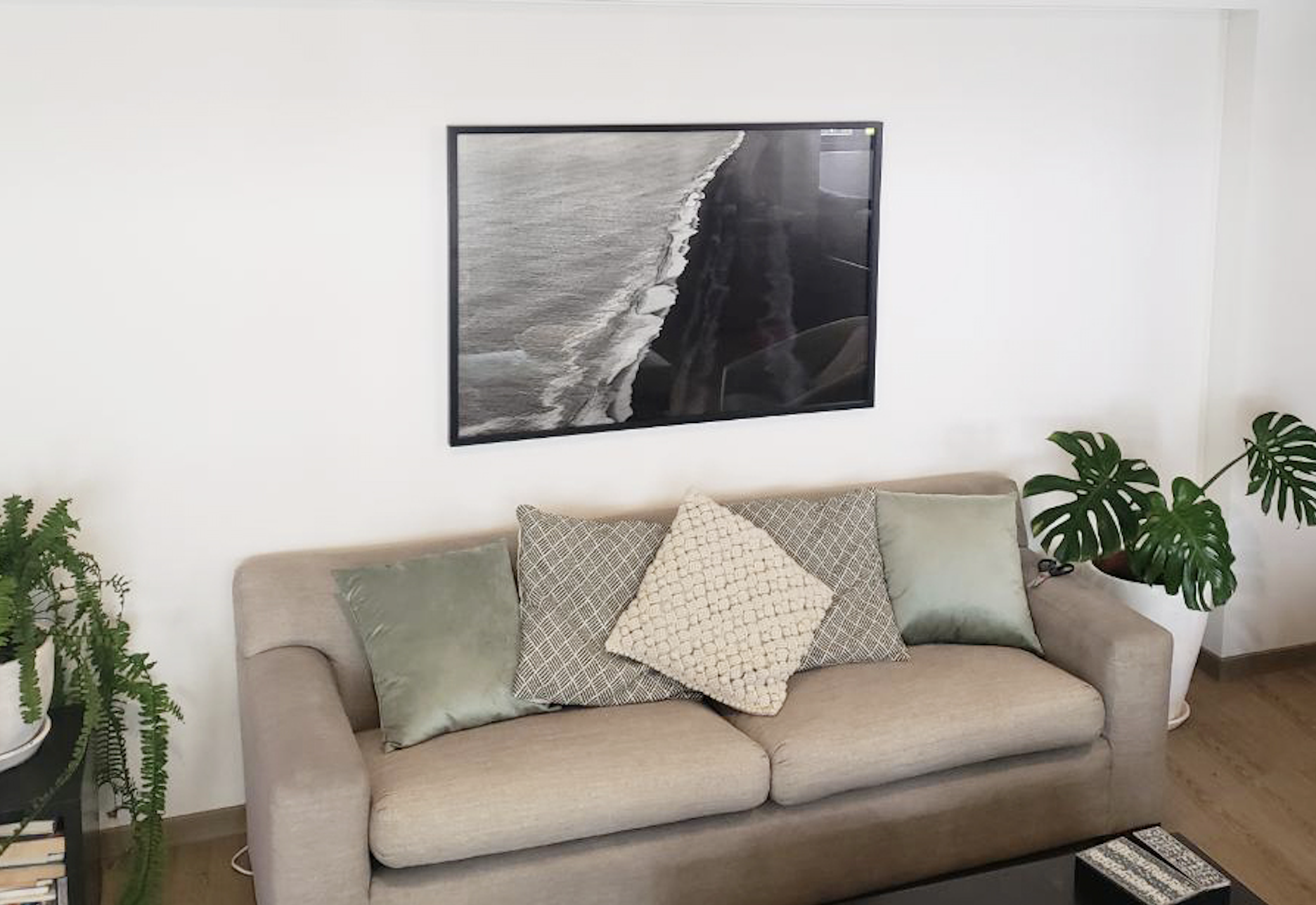 Sala de estar con sofá beige, cojines decorativos, pintura de playa en la pared y plantas verdes a la derecha y izquierda.