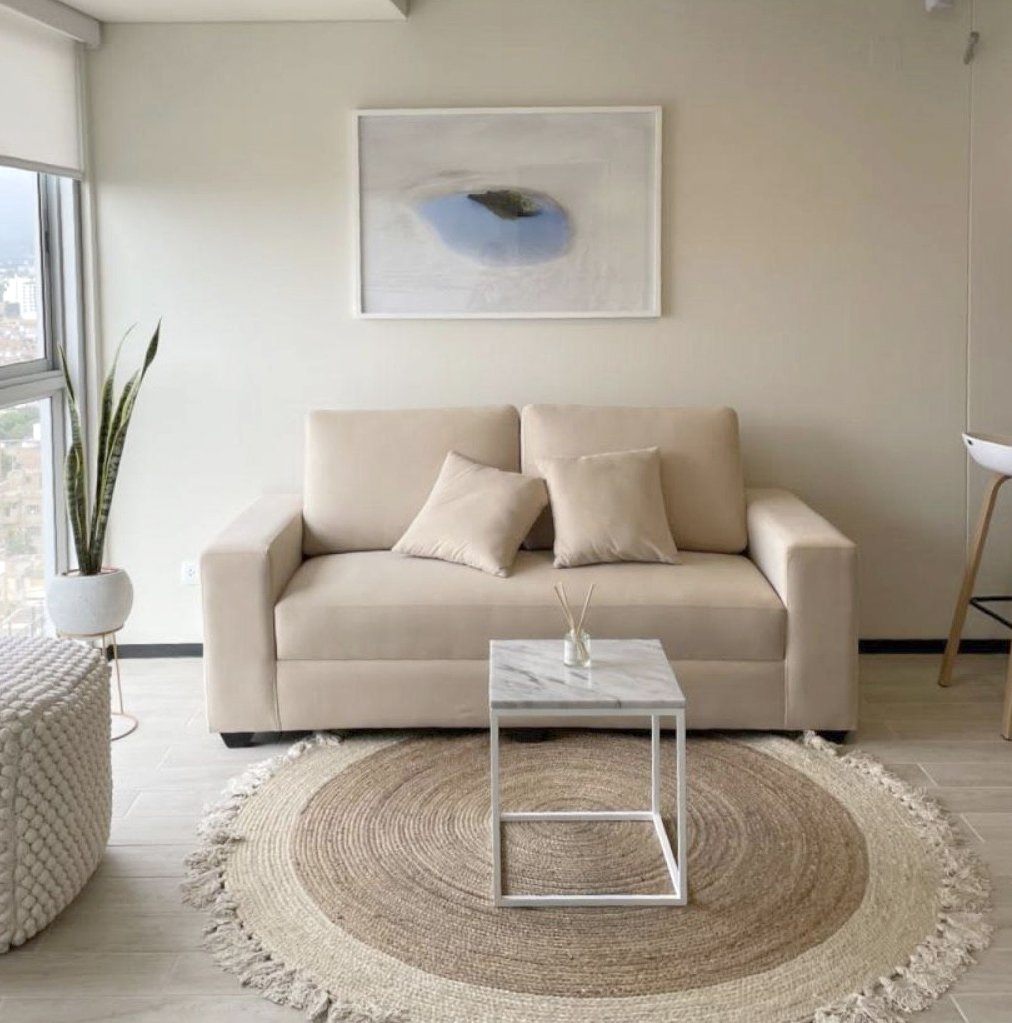 Sala de estar con sillón beige, mesa de café de mármol, alfombra redonda tejida, cuadro abstracto en la pared, planta en maceta y ventana grande con vista a la ciudad.
