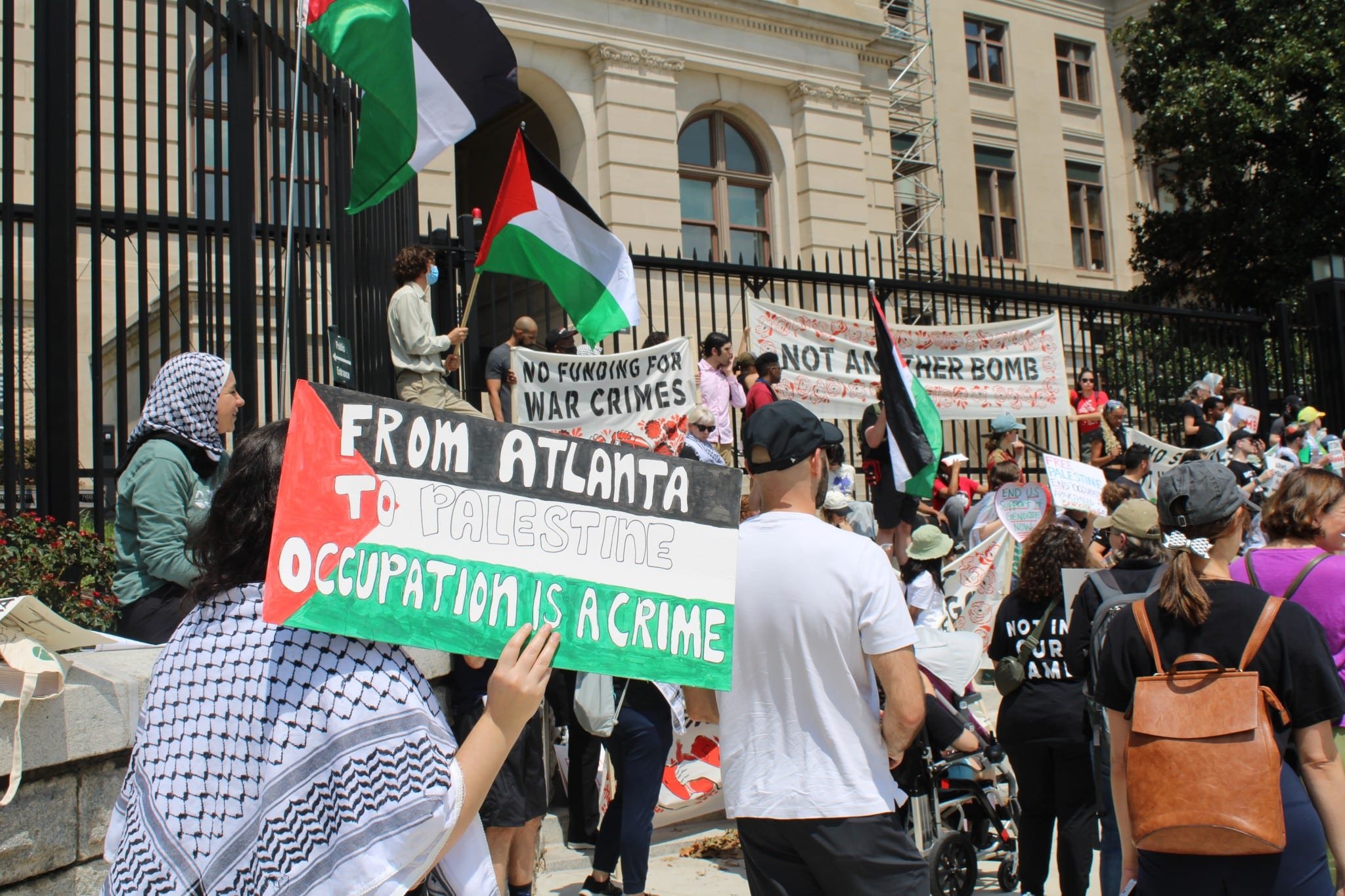 A diverse group of people participating in a protest or rally outdoors, holding signs and flags. The signs include messages such as 'No funding for war crimes' and 'From Atlanta to Palestine, Occupation is a crime.' Some protestors are wearing masks, and one person is holding a Palestinian flag. The scene is set against a building with windows and a large black fence.