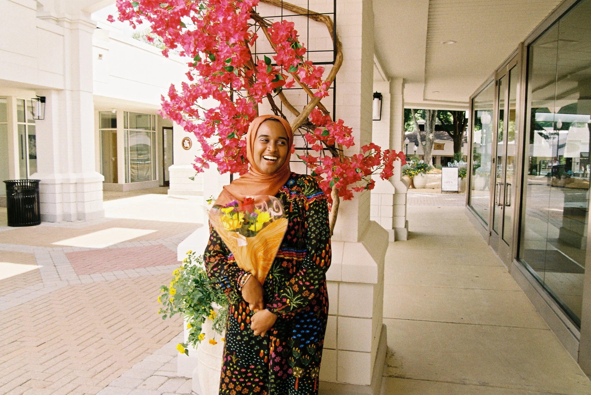 A smiling woman wearing a peach-colored headscarf and a dark dress with colorful floral patterns. She is holding a bouquet of yellow and purple flowers and standing next to a pink flowering tree in an outdoor shopping area.