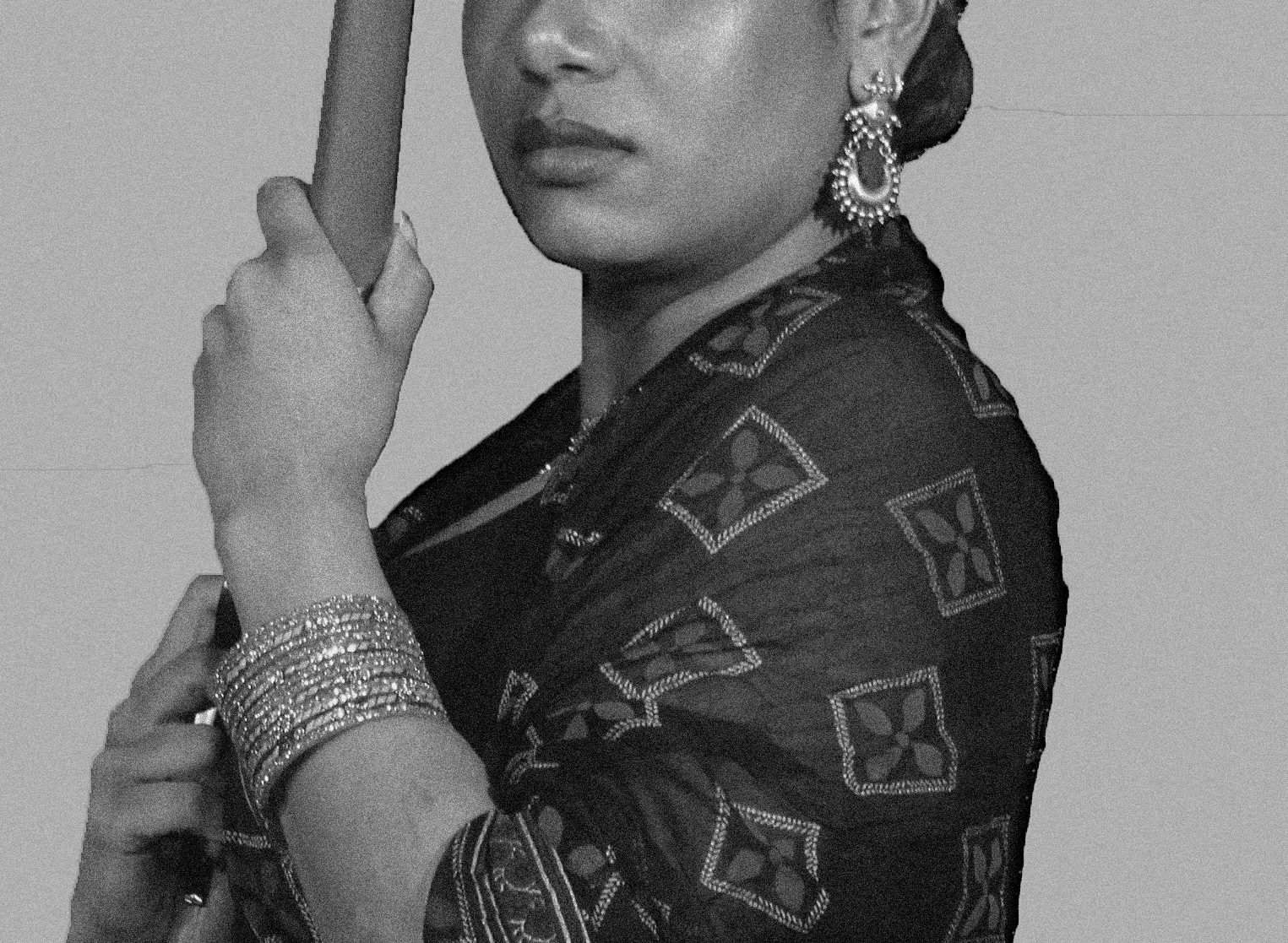 A woman dressed in traditional attire with jewelry, her hand is raised near her face, and she is holding an object, possibly a stick or a rod, against a plain background.