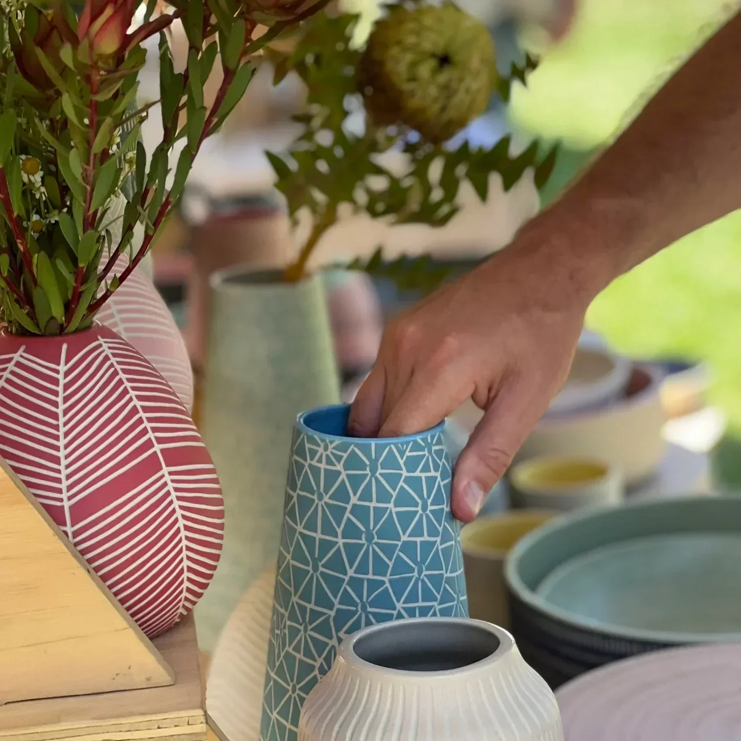 Close-up of a person's hand reaching into a blue geometric-patterned vase with plants on display, surrounded by colorful bowls and plants.