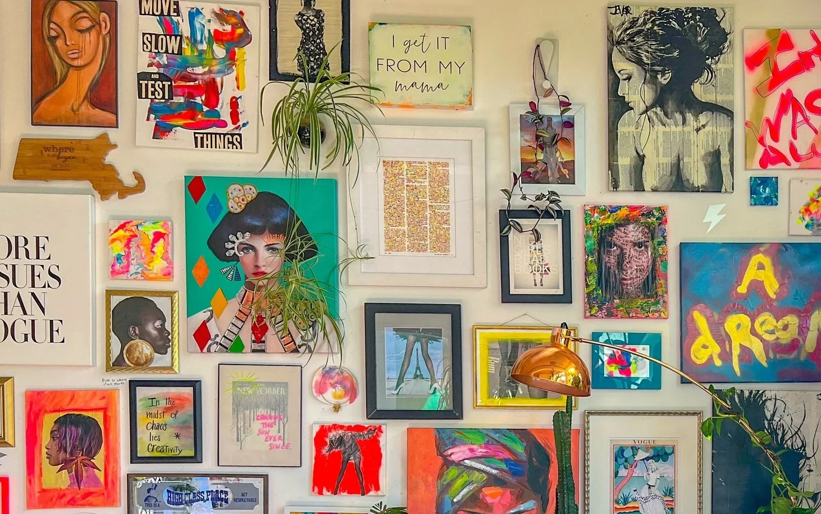 A colorful gallery wall featuring various framed artworks, photographs, and motivational quotes, with some plants and a copper-colored desk lamp.