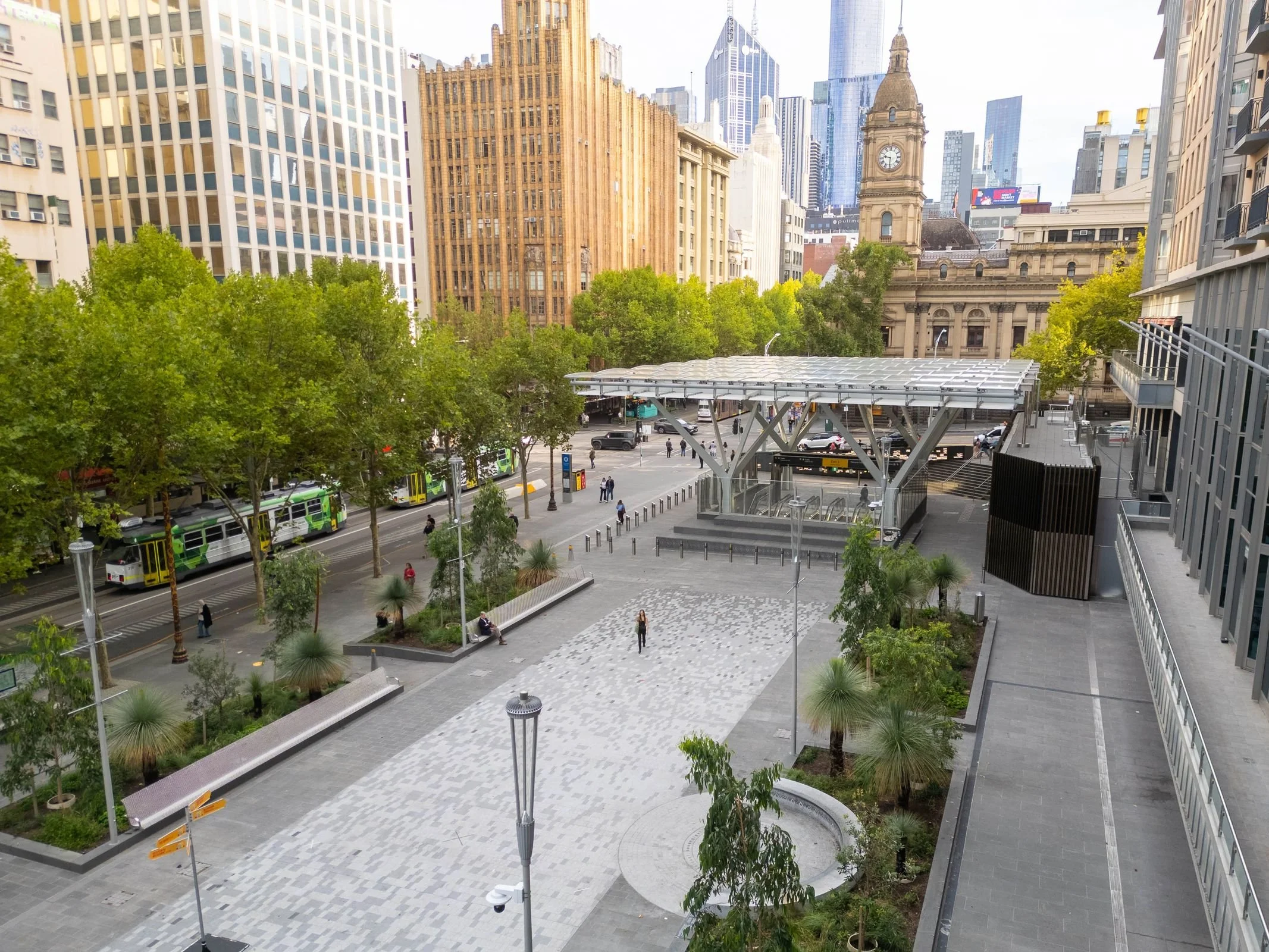 Cityscape view with trees in the foreground and tall buildings including a clock tower in the background, along with a public transportation station and a street with buses and pedestrians.