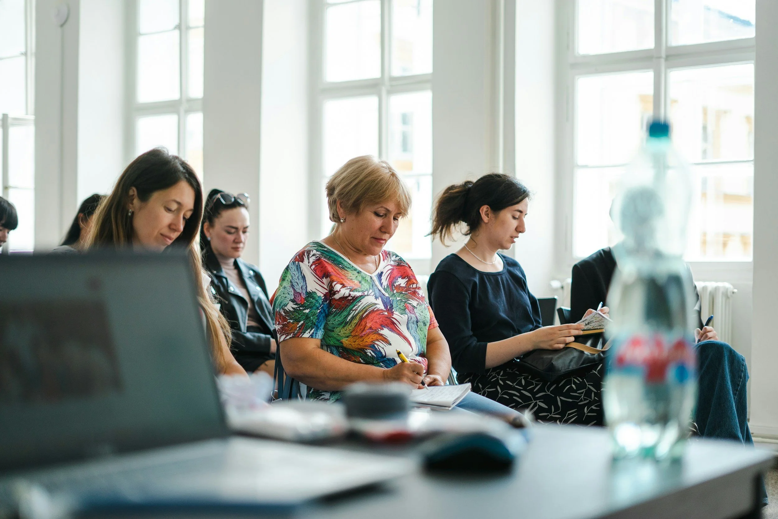 A group of women sitting in a bright room with large windows, engaging in a workshop or seminar, with some taking notes and focused on the session.