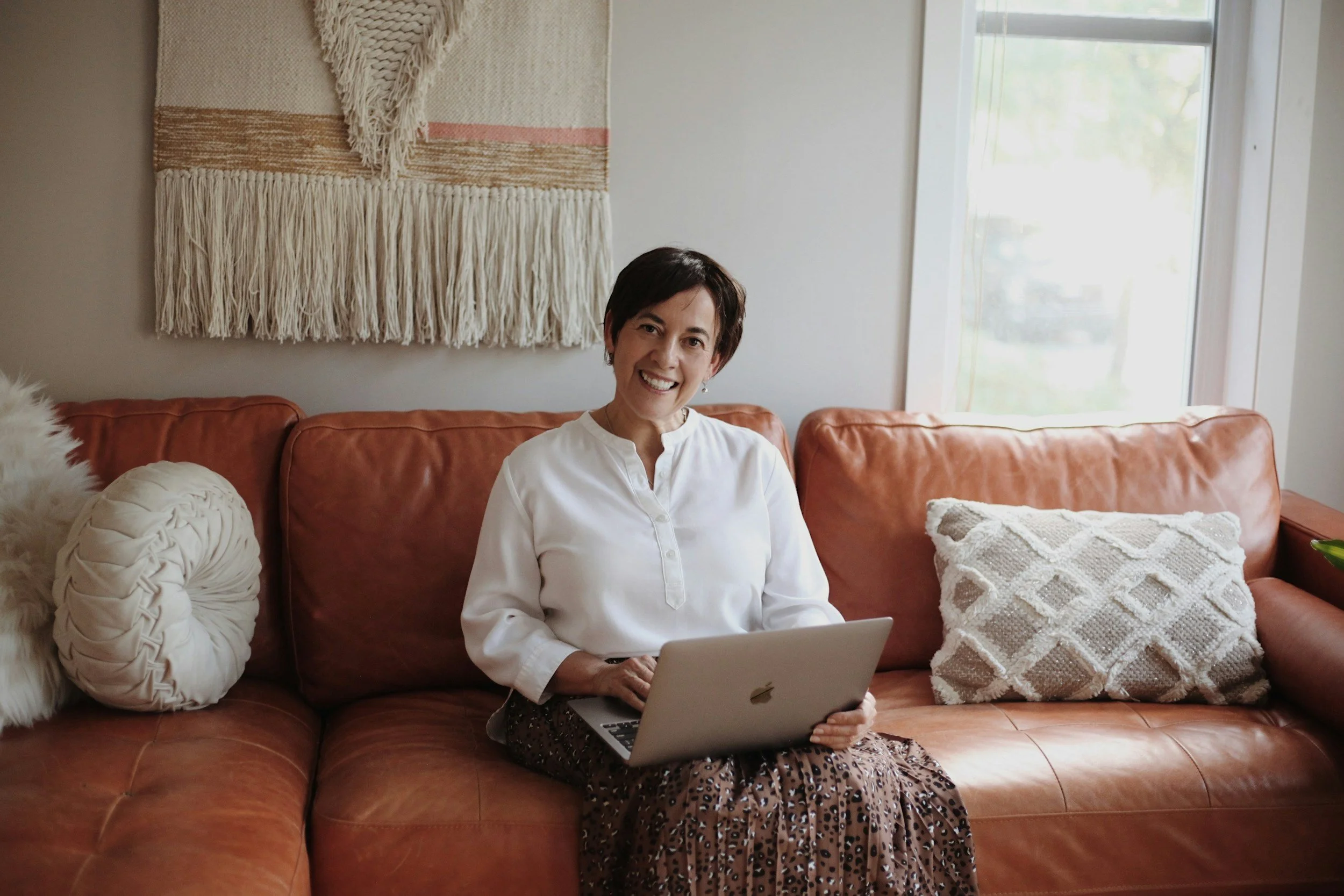 A woman with short dark hair, wearing a white blouse and patterned skirt, sitting on a brown leather couch with decorative pillows, using a silver laptop in a bright living room with a window and wall tapestry.