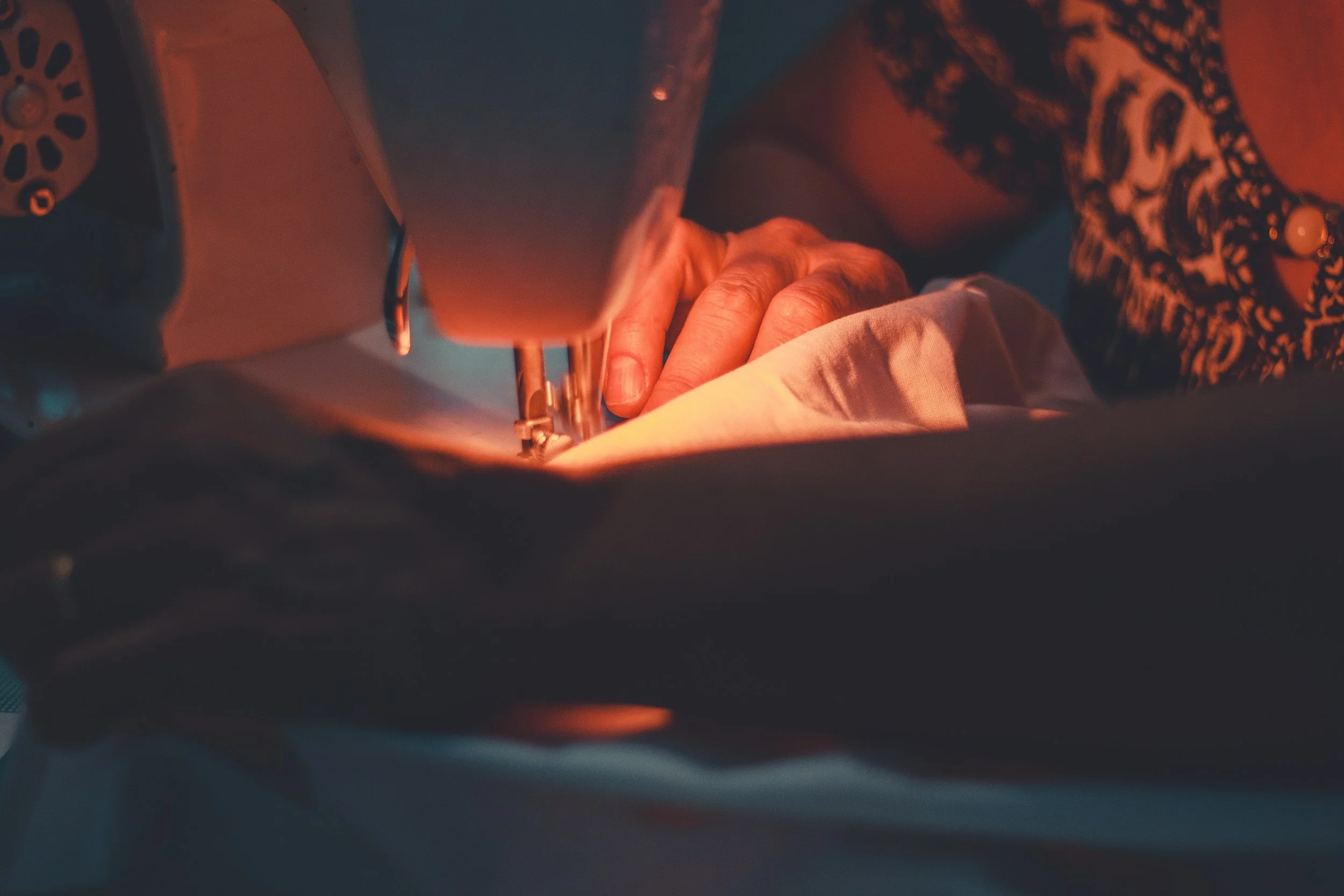 Close-up of a person sewing fabric on a sewing machine, with their hand guiding the fabric through the machine, illuminated by warm light.
