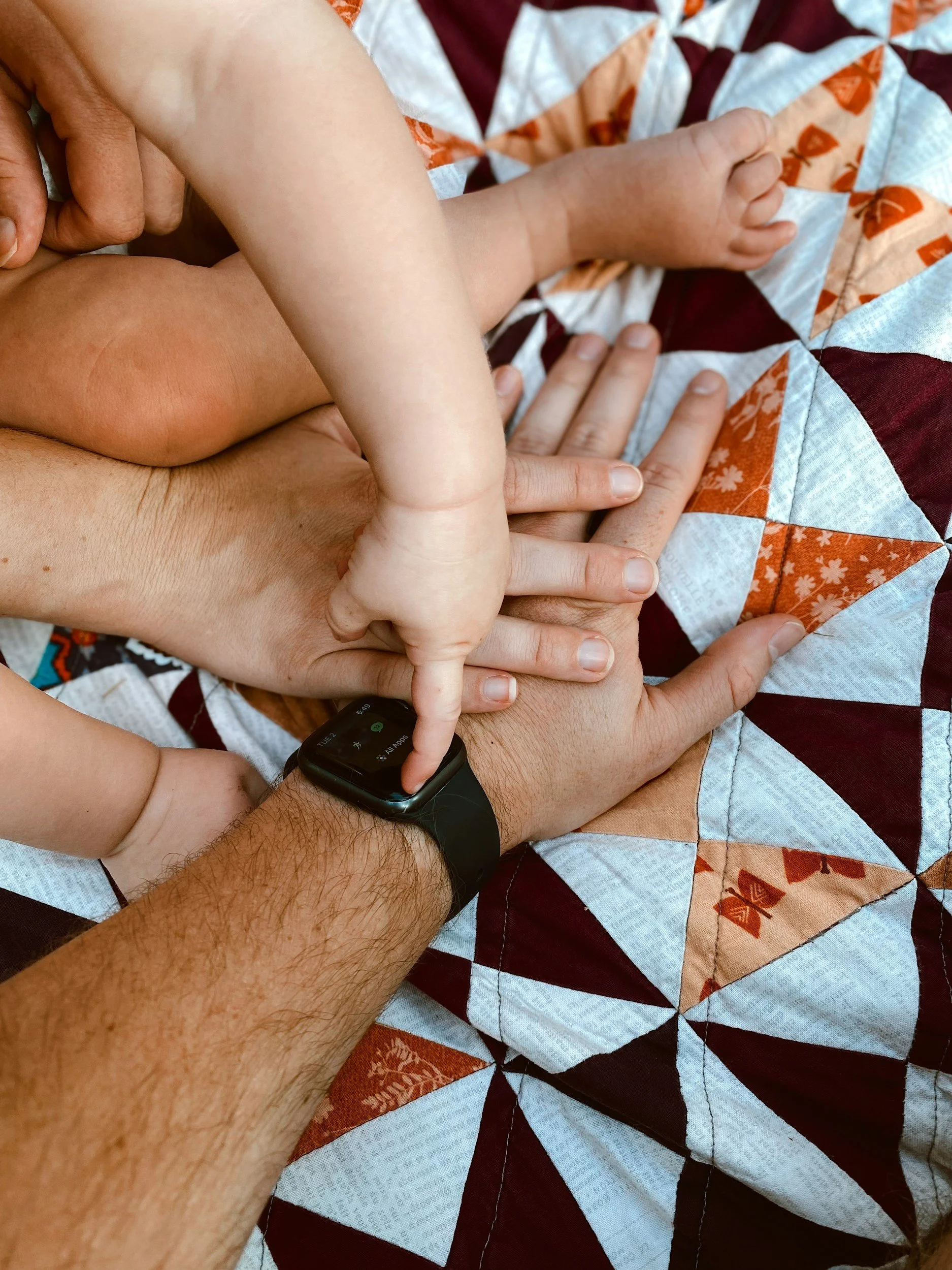 Close-up of hands, including an adult's and a child's, laying on a quilted bedspread with geometric patterns in red, white, orange, and brown. The adult is wearing a black smartwatch.