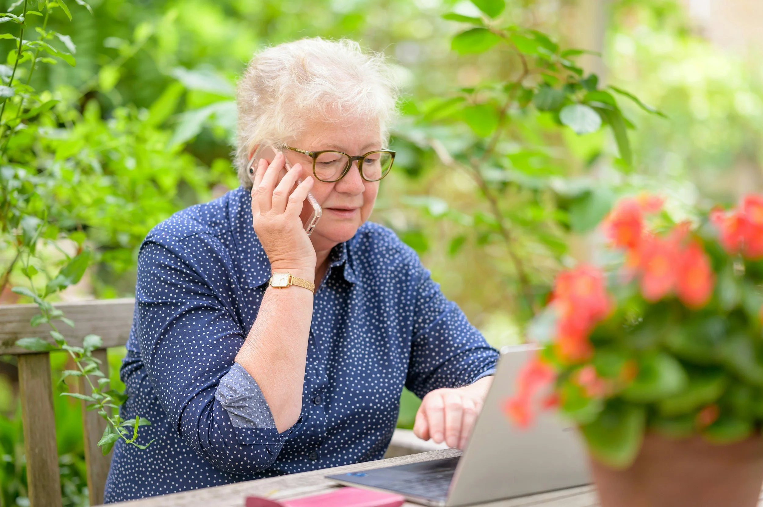 An elderly woman with white hair, glasses, and a blue polka-dotted shirt talking on the phone while working on a laptop outdoors in a lush, green garden.