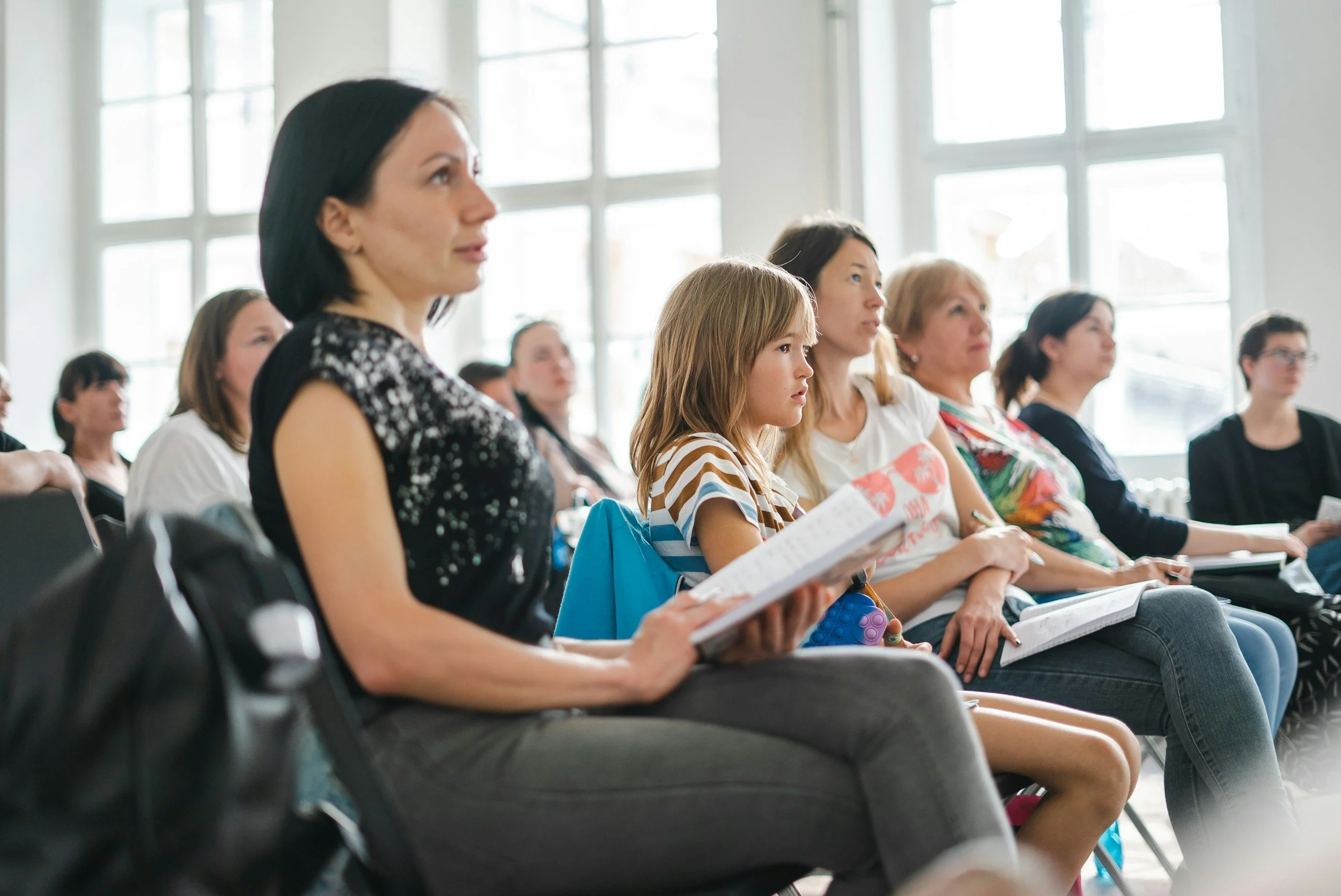 Group of diverse women and girls sitting in a bright room, attending a lecture or presentation, some taking notes.