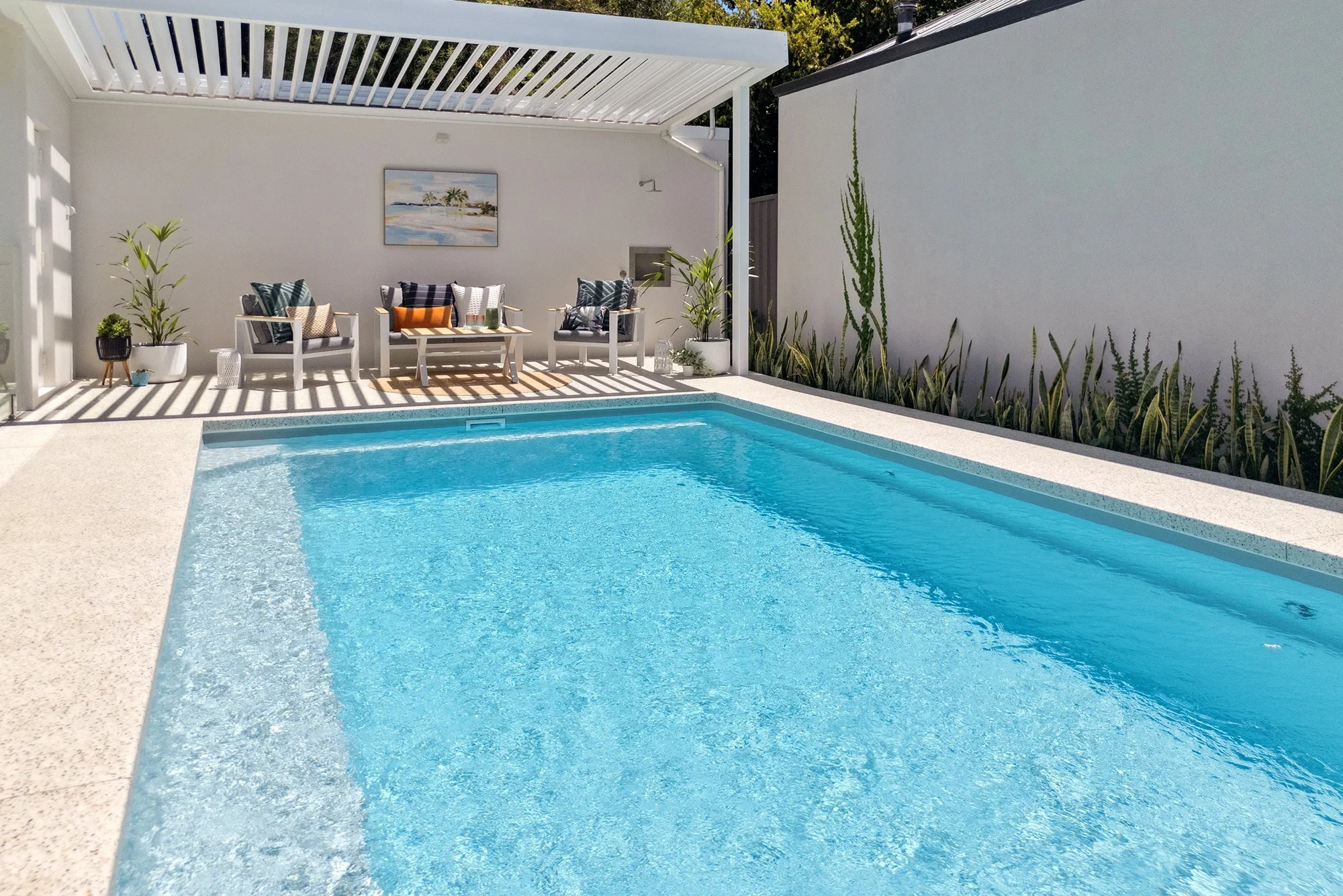Light blue swimming pool set in marble deck. Deck chairs in background against white wall.
