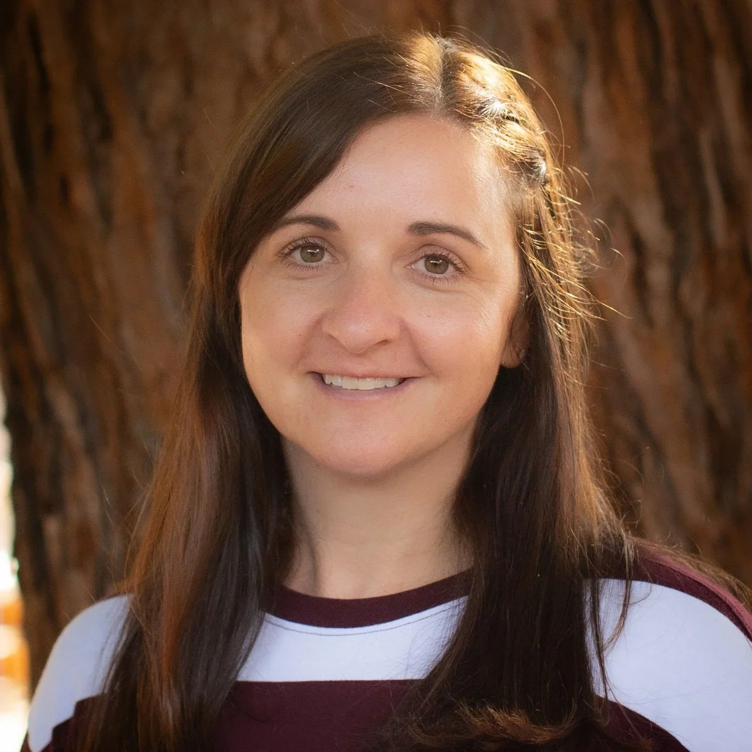 Close-up of a smiling woman with long brown hair, wearing a white and maroon shirt, standing outdoors with a tree trunk in the background.