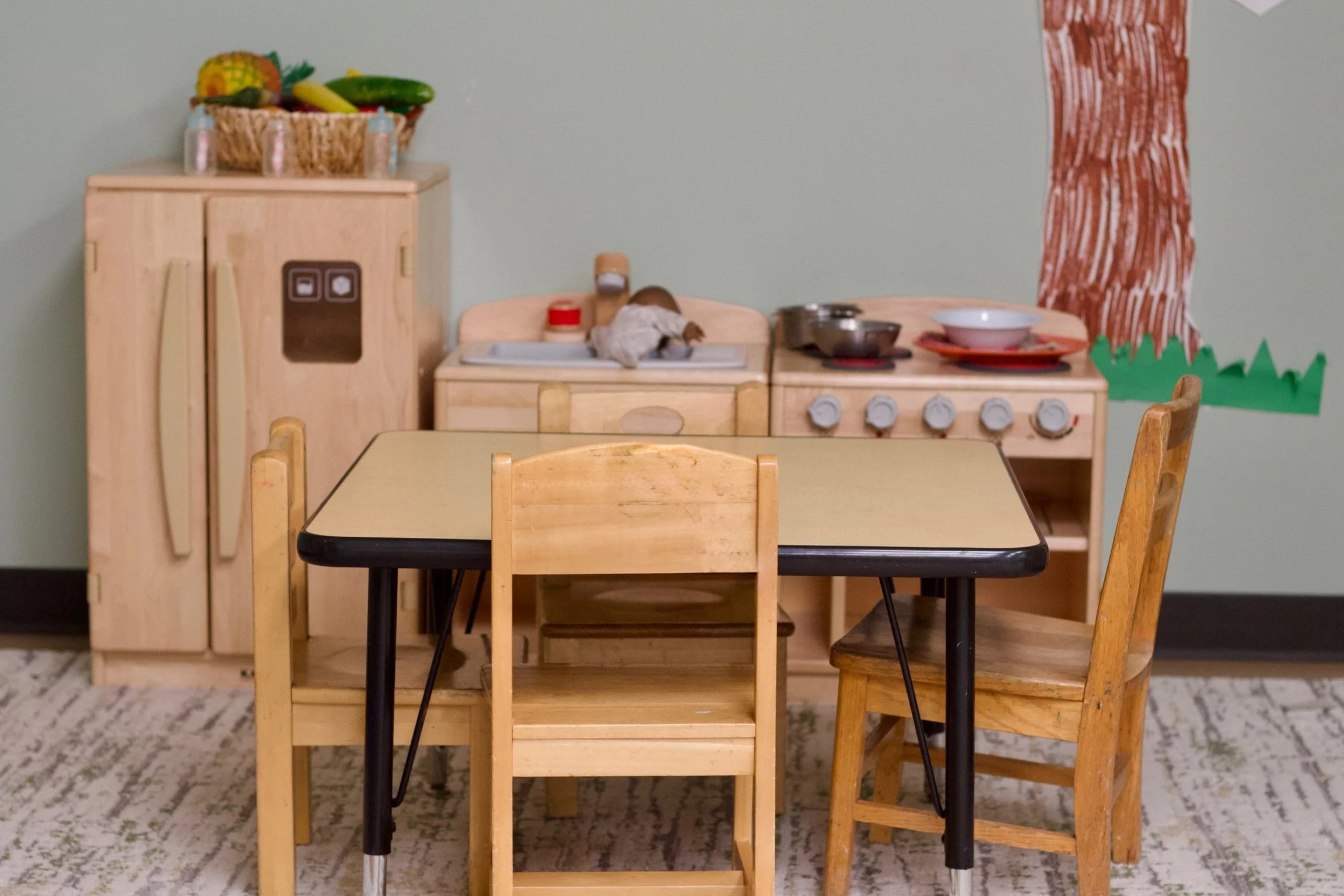 A small wooden play kitchen with a basket of toy fruits and vegetables on top, a mouse figurine on the stove, and a table with chairs in front. The wall behind has a red and green paper decoration.