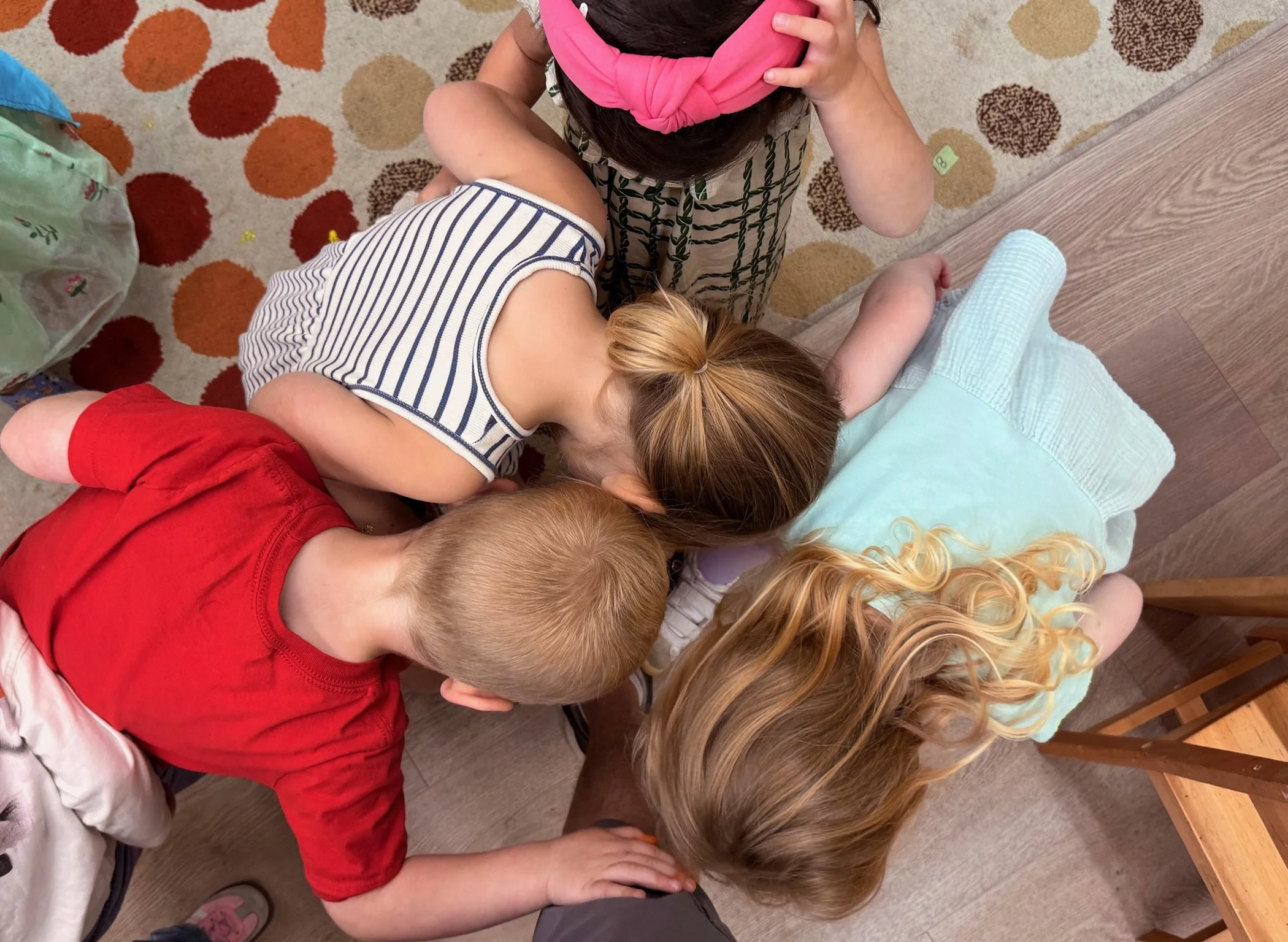 Children gathered closely in a circle, leaning in and focusing on something in the center, on a wooden floor with a patterned rug.
