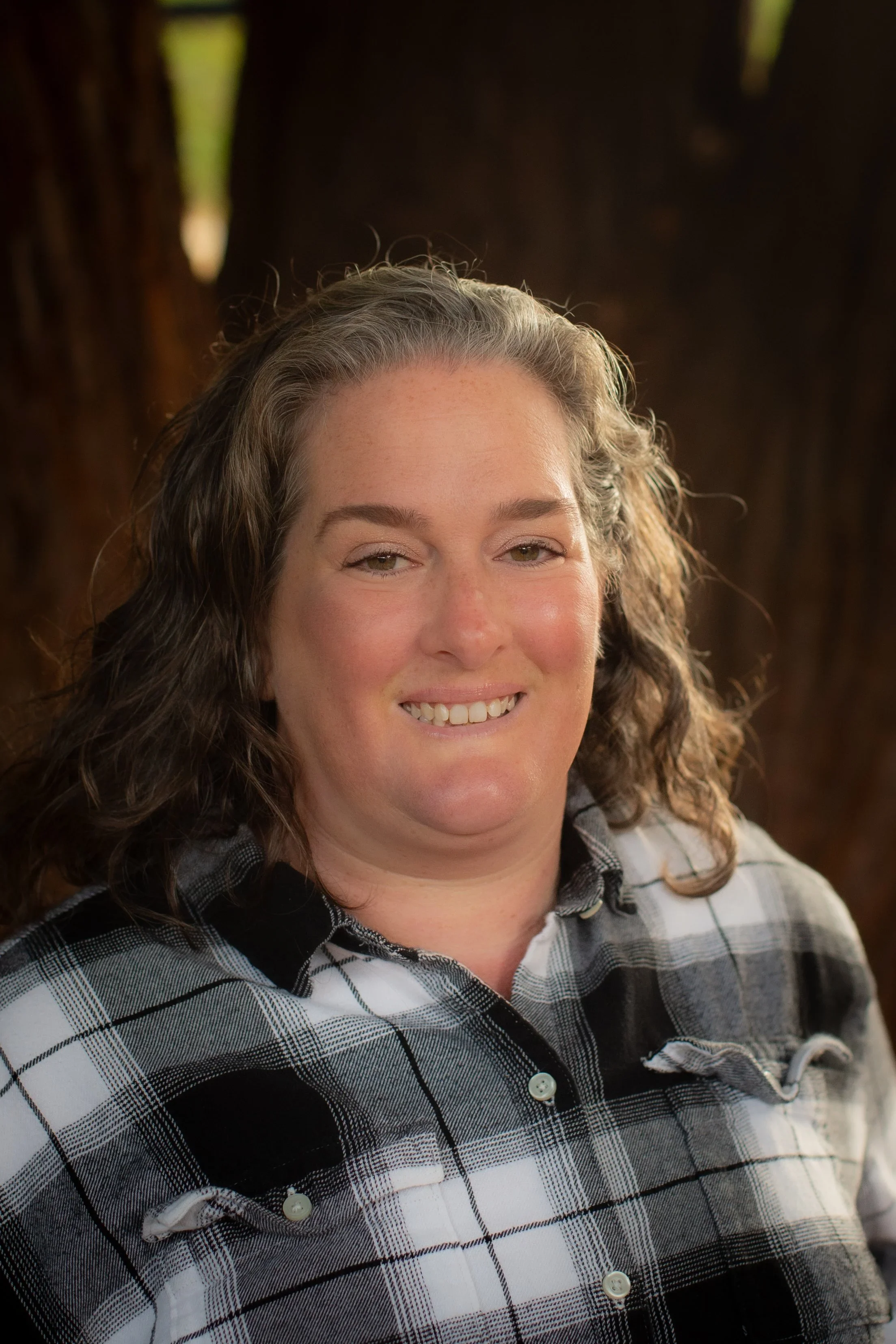 A woman with wavy brown hair, smiling, wearing a black, white, and gray plaid shirt with buttons, in front of a dark brown background.