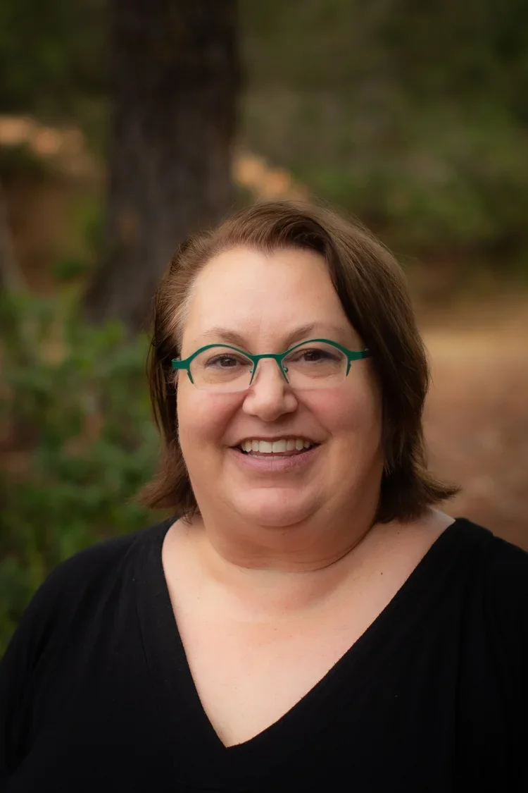 A smiling woman with glasses and short brown hair, wearing a black top, standing outdoors with trees and greenery in the background.