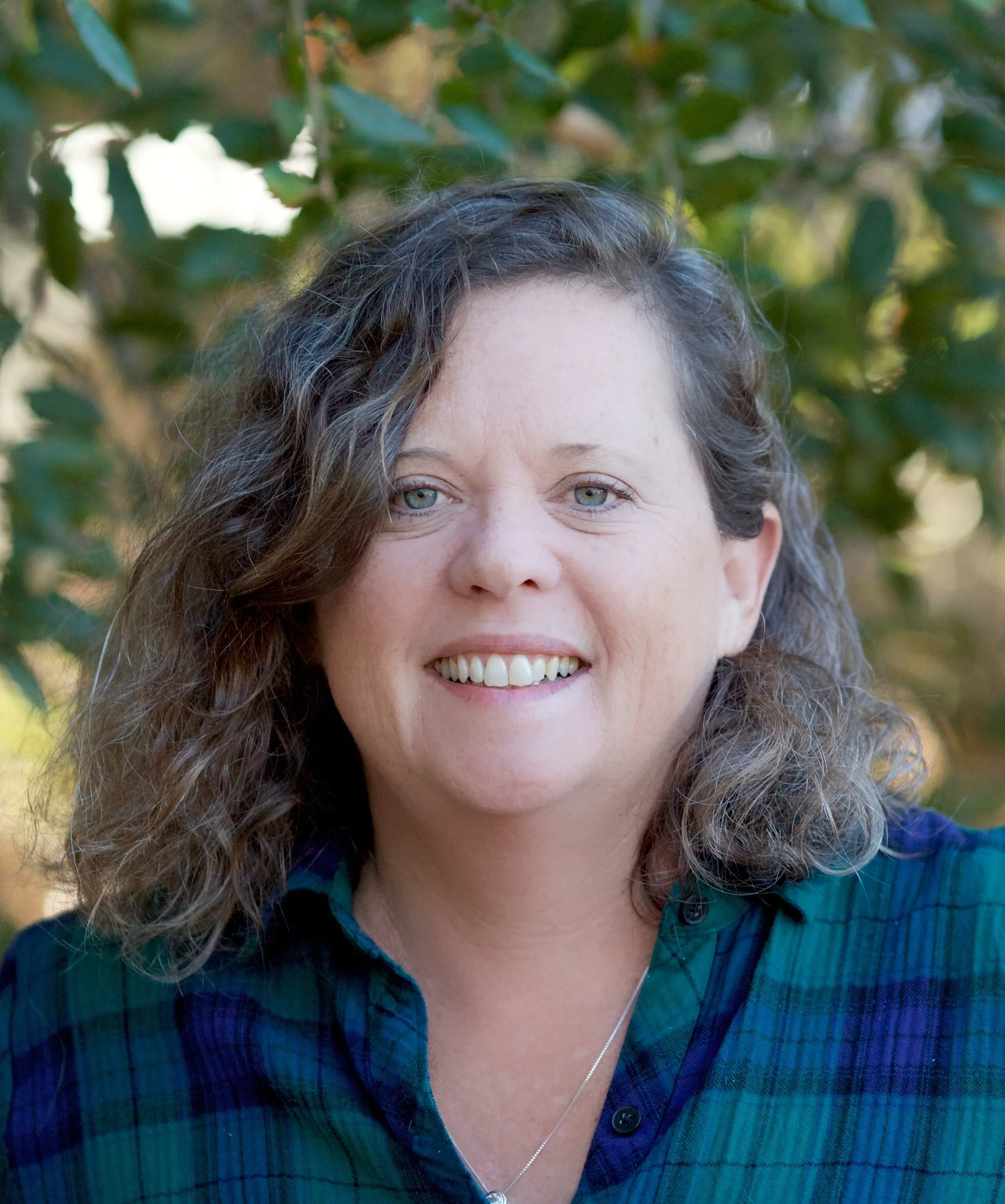 Close-up of a smiling woman with curly brown hair, wearing a blue plaid shirt and a necklace, standing outdoors with greenery in the background.
