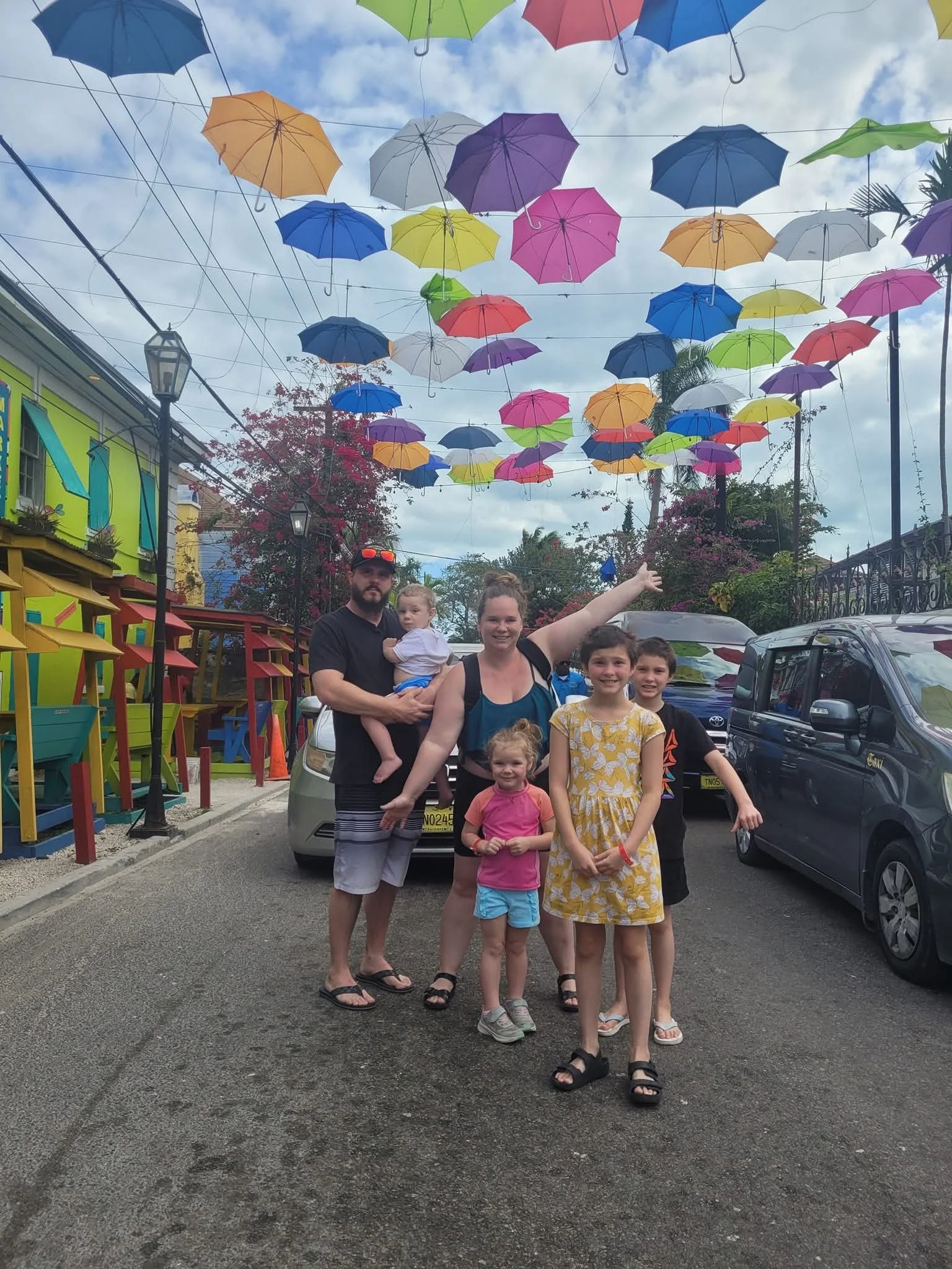 A family of seven standing on a street, with colorful umbrellas hanging above them in the sky, surrounded by buildings and parked cars.