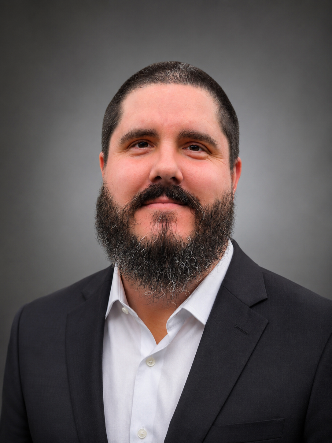 A man with a beard and short dark hair wearing a black suit and white shirt against a gray background.