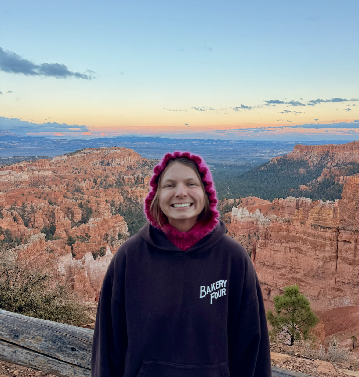 Smiling woman in black hoodie with pink hat standing in front of canyon landscape during sunset.